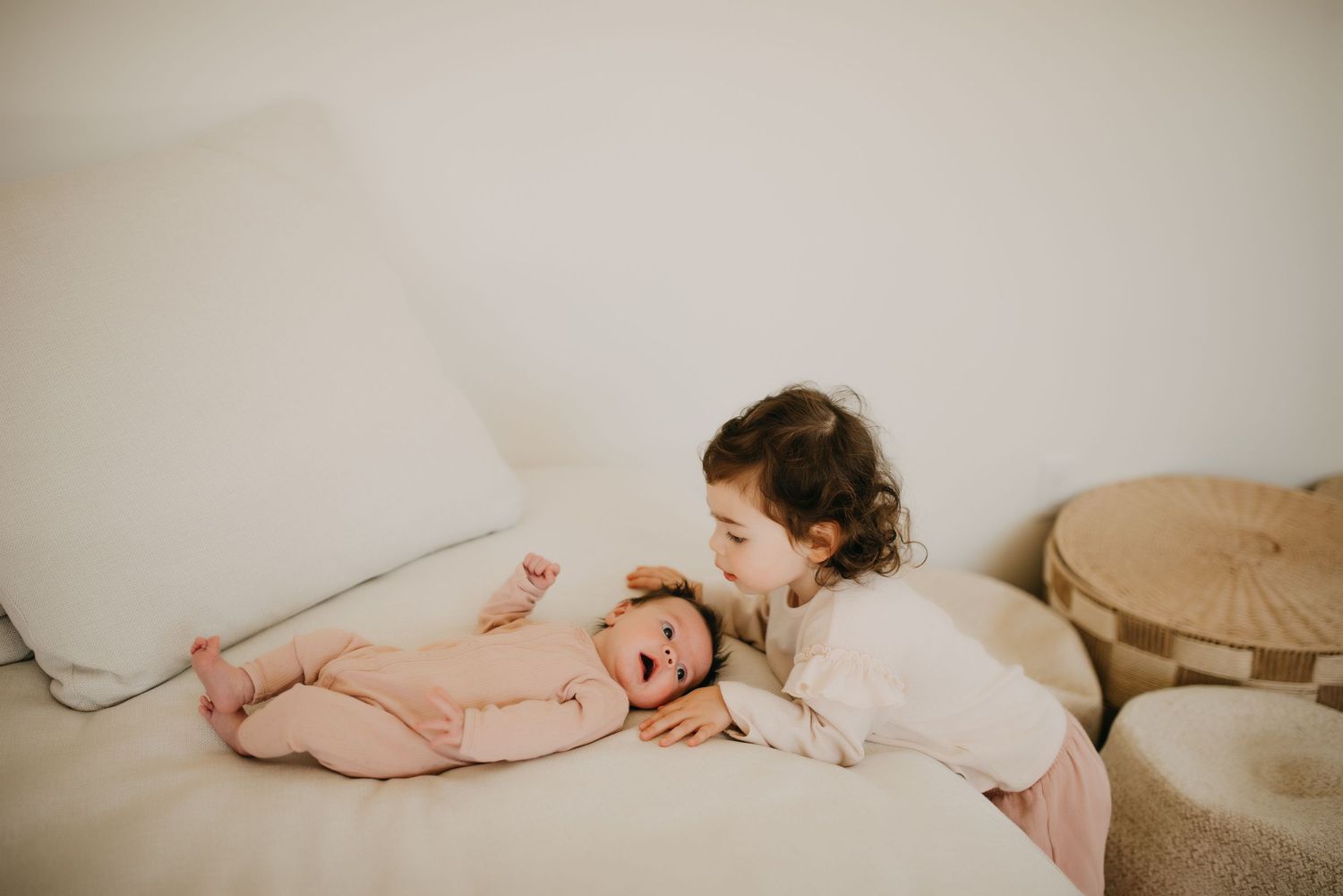 Curly-haired toddler leaning in to kiss newborn sibling lying on white bed.