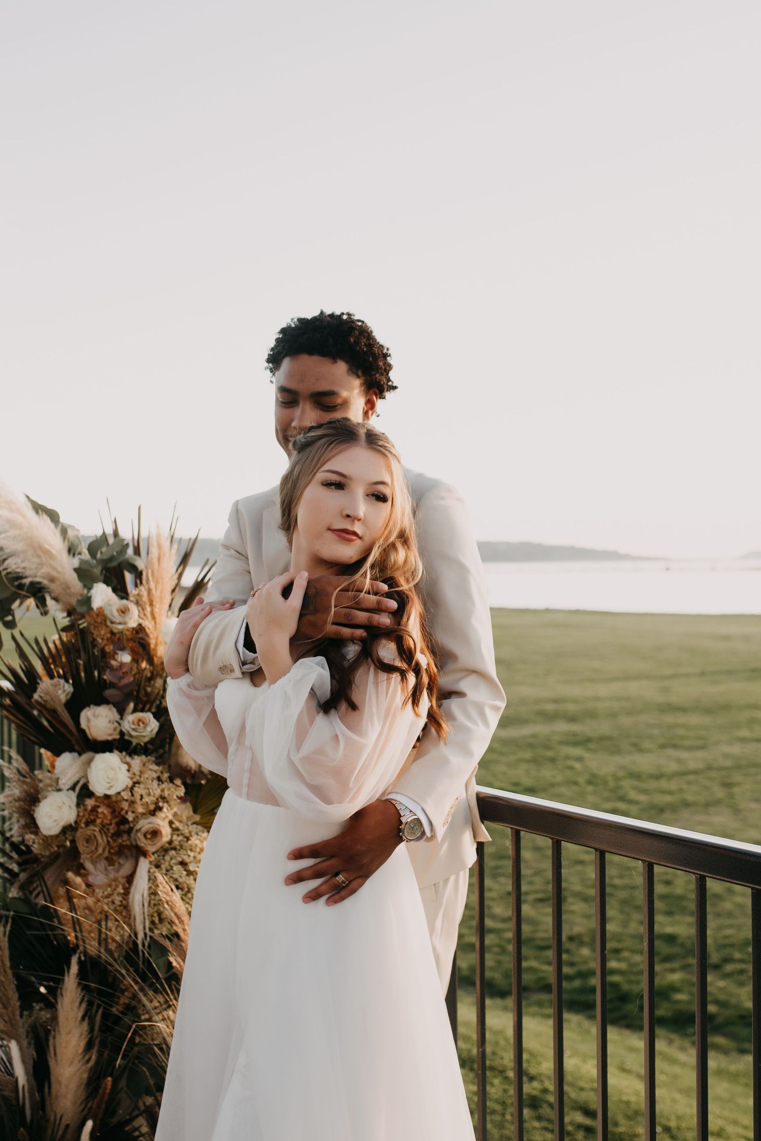 Groom in a beige suit embracing bride in white dress on a balcony overlooking a green field.