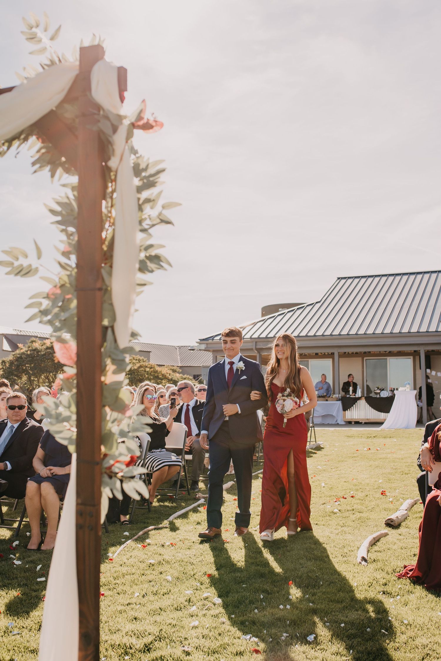 Groomsman escorting bridesmaid in red dress down outdoor wedding ceremony aisle on sunny day.