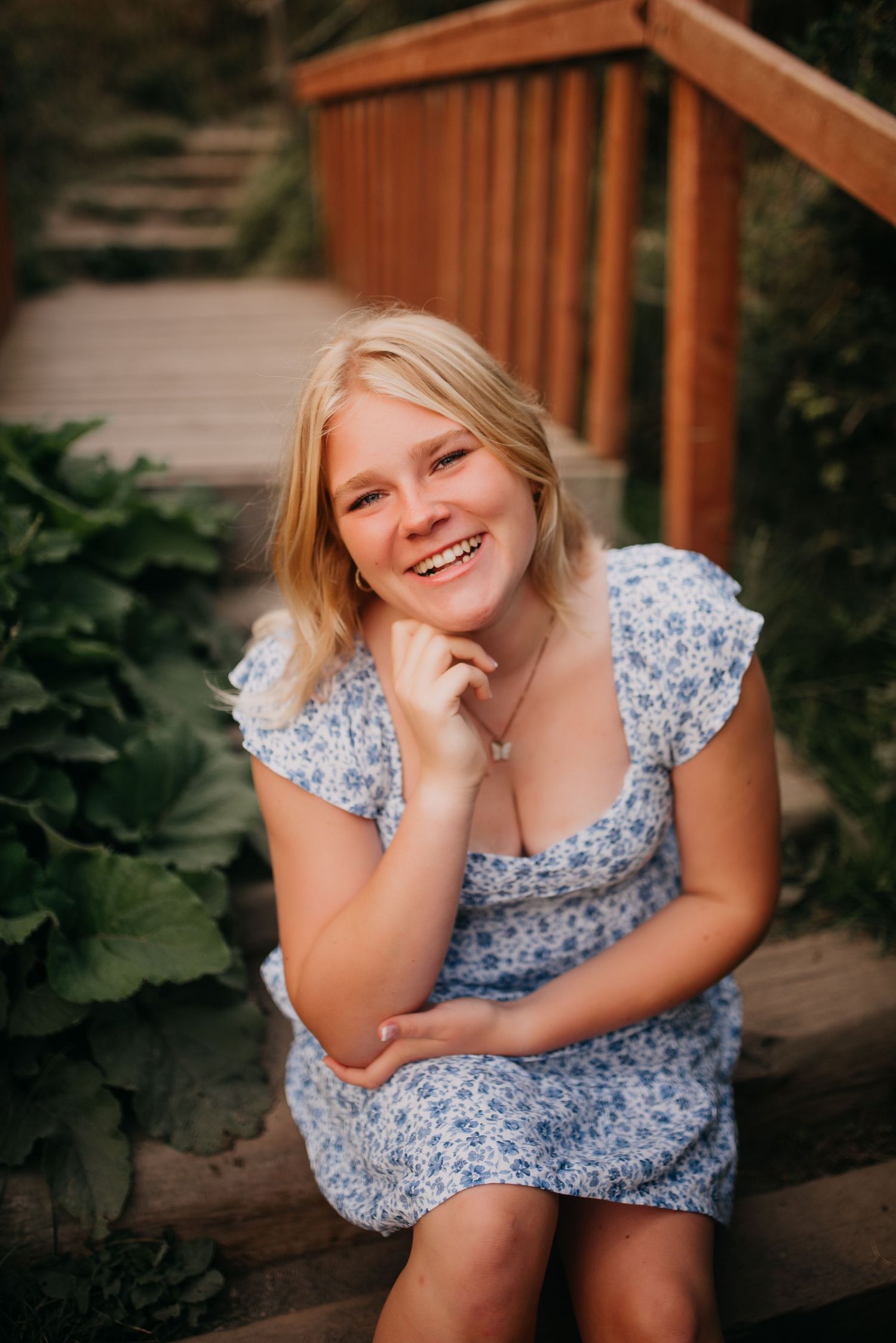 Smiling blonde young woman in blue floral dress sits on wooden garden steps surrounded by greenery.