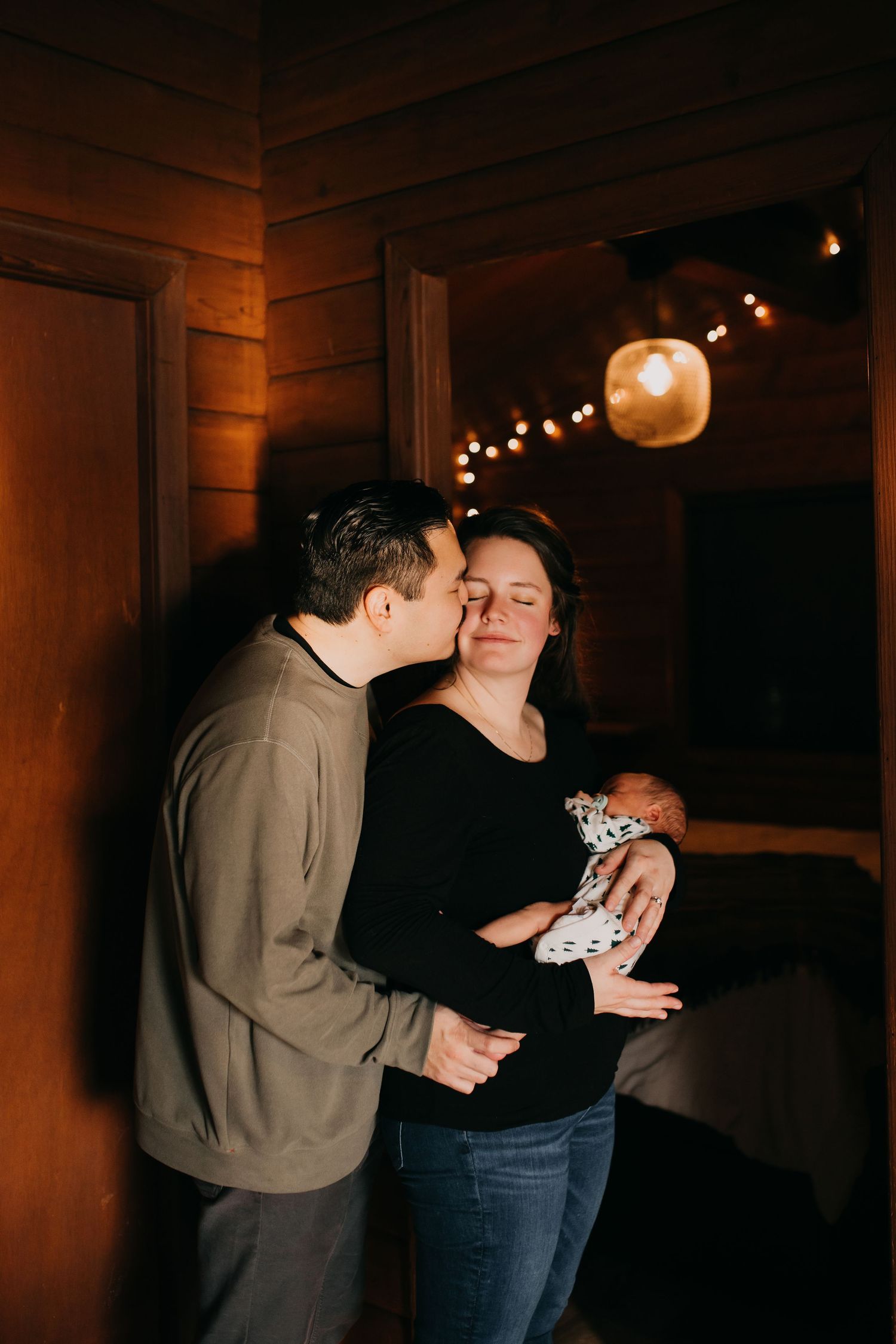 Loving couple with newborn baby sharing a kiss in warmly lit wooden interior.