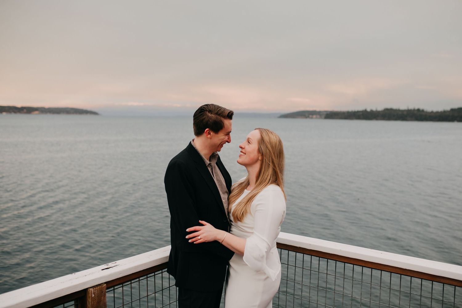 Romantic couple nearly kissing on waterfront deck at sunset with calm ocean behind them.