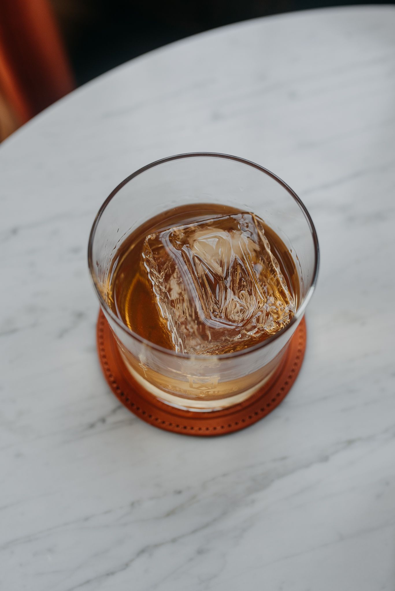 Top view of a whiskey cocktail with large ice cube in a rocks glass on an orange leather coaster.