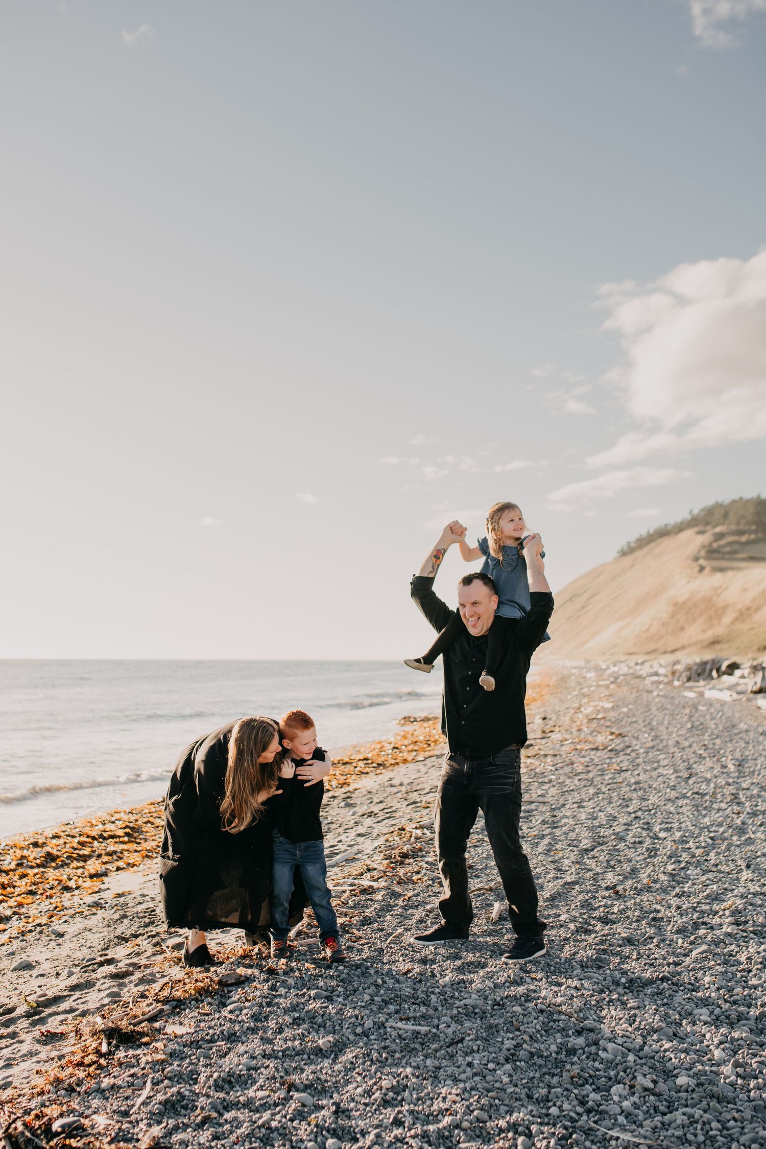 Family of four playing on rocky Pacific Northwest beach at golden hour with coastal bluffs in background.