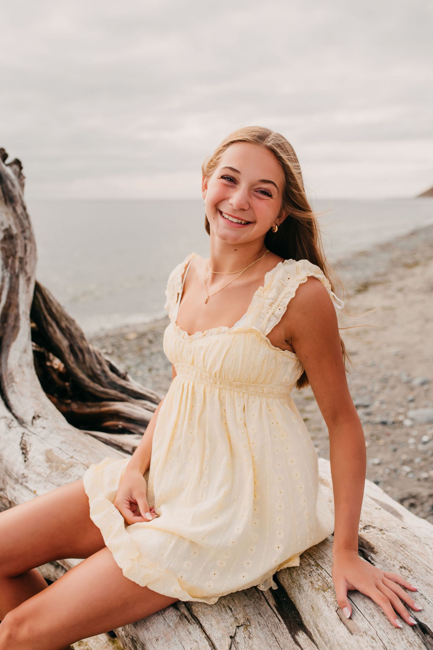 Smiling teenage girl in yellow sundress sits on driftwood at a rocky beach.