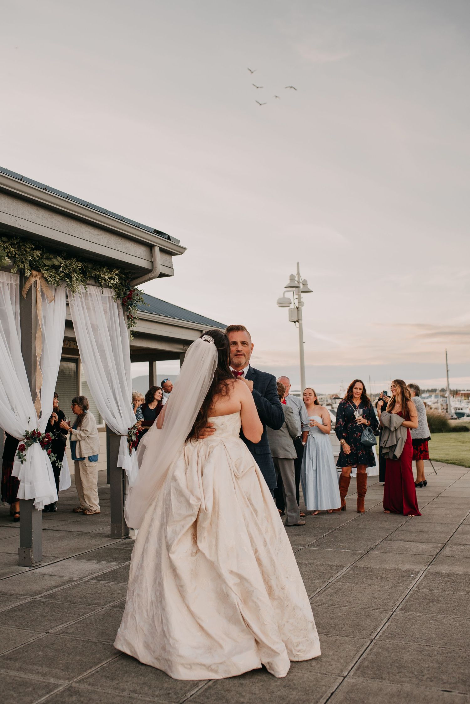 Bride and groom share first dance outdoors at waterfront wedding venue at dusk with guests watching.