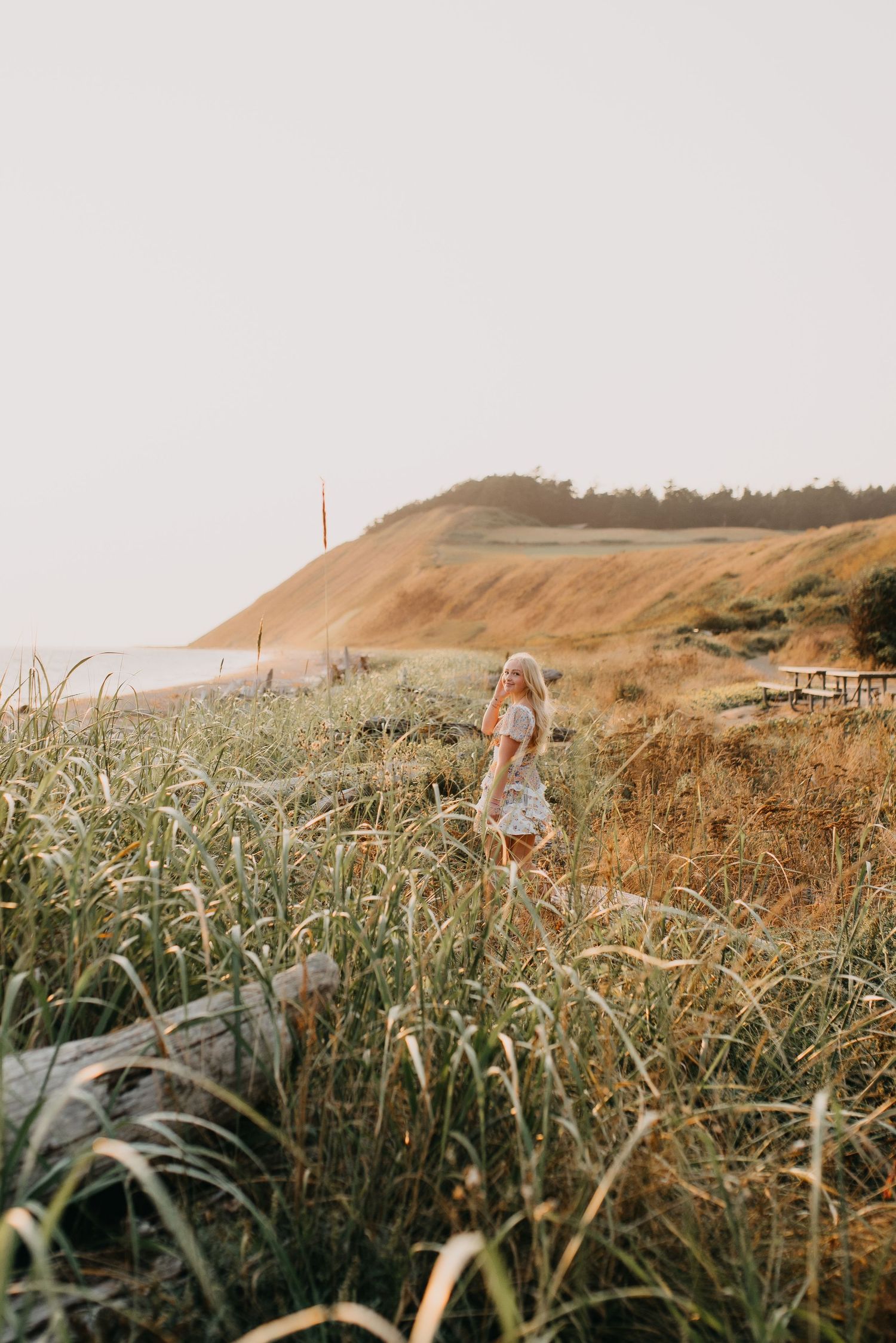 Blonde woman in floral dress walks through tall beach grass with sandy dunes and ocean in the background.