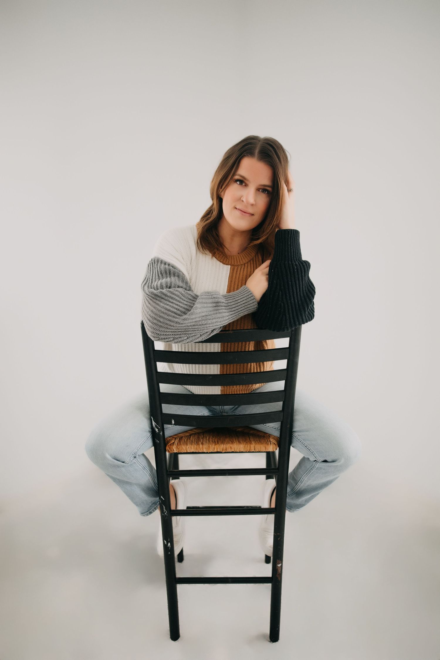 Young woman in colorblock sweater sitting backwards on wooden chair against white background.
