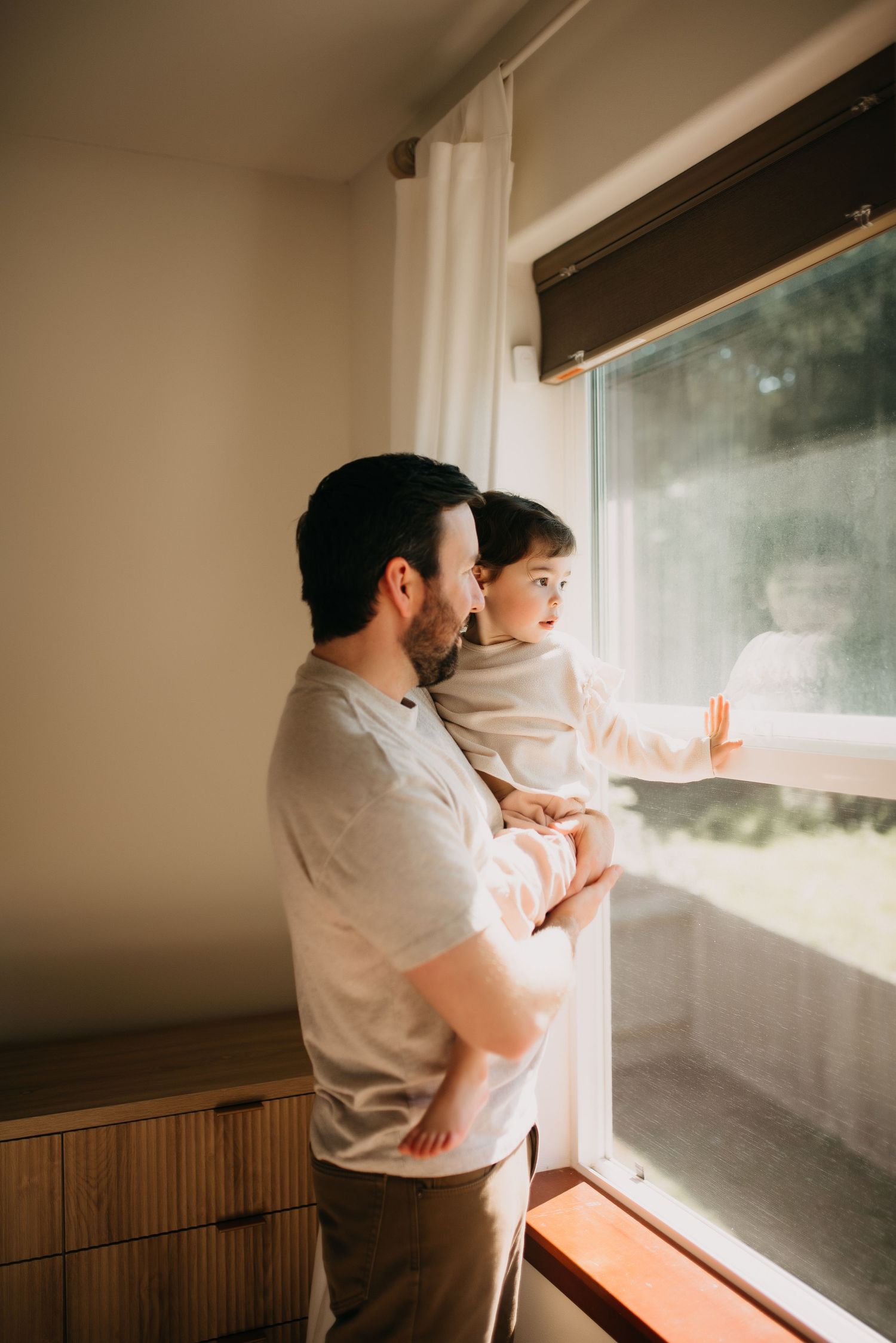 Father holding young toddler while both look out a sunlit window in a warmly lit modern home.