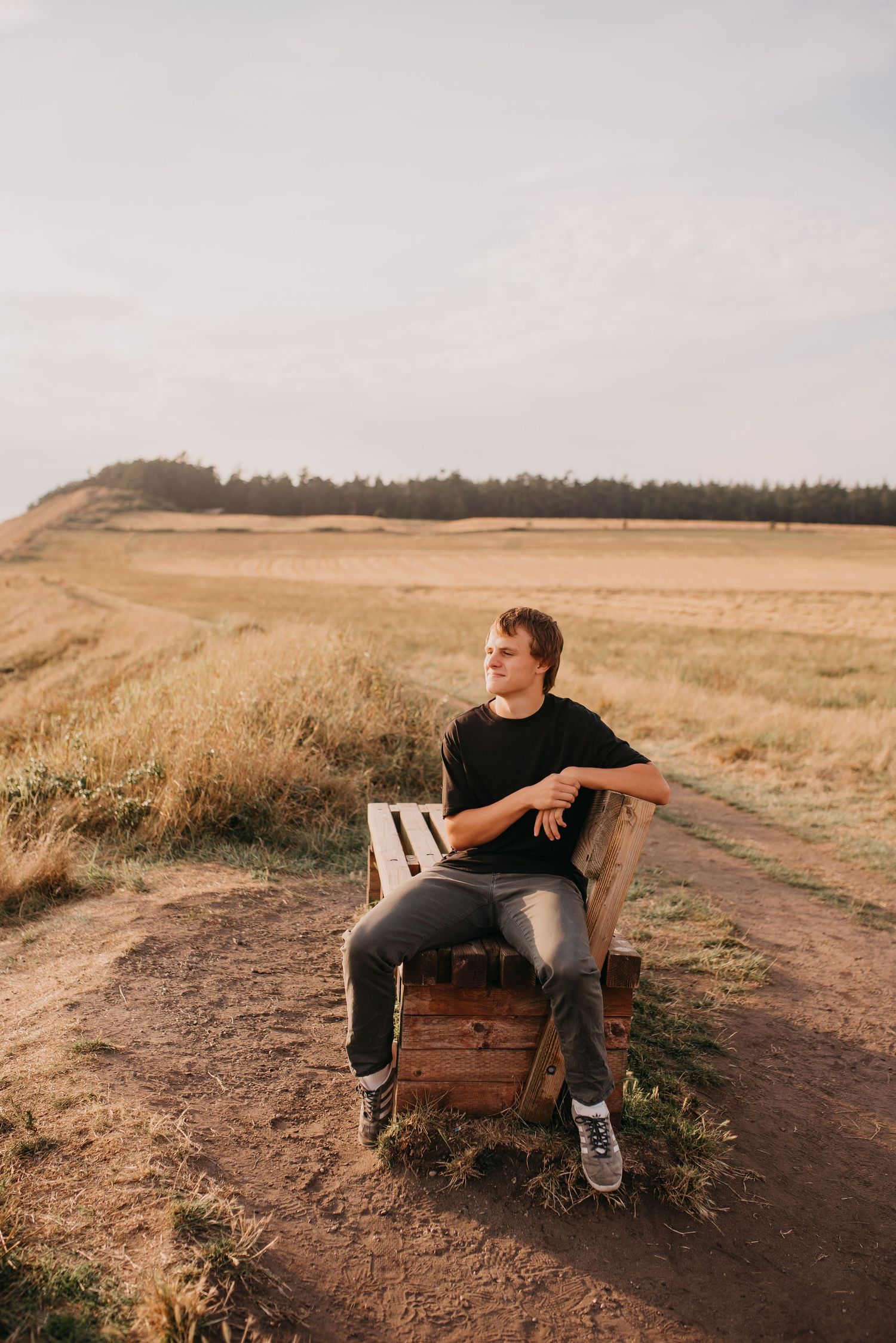 Young man in black t-shirt sits on wooden crate beside rural dirt path in golden open farmland.