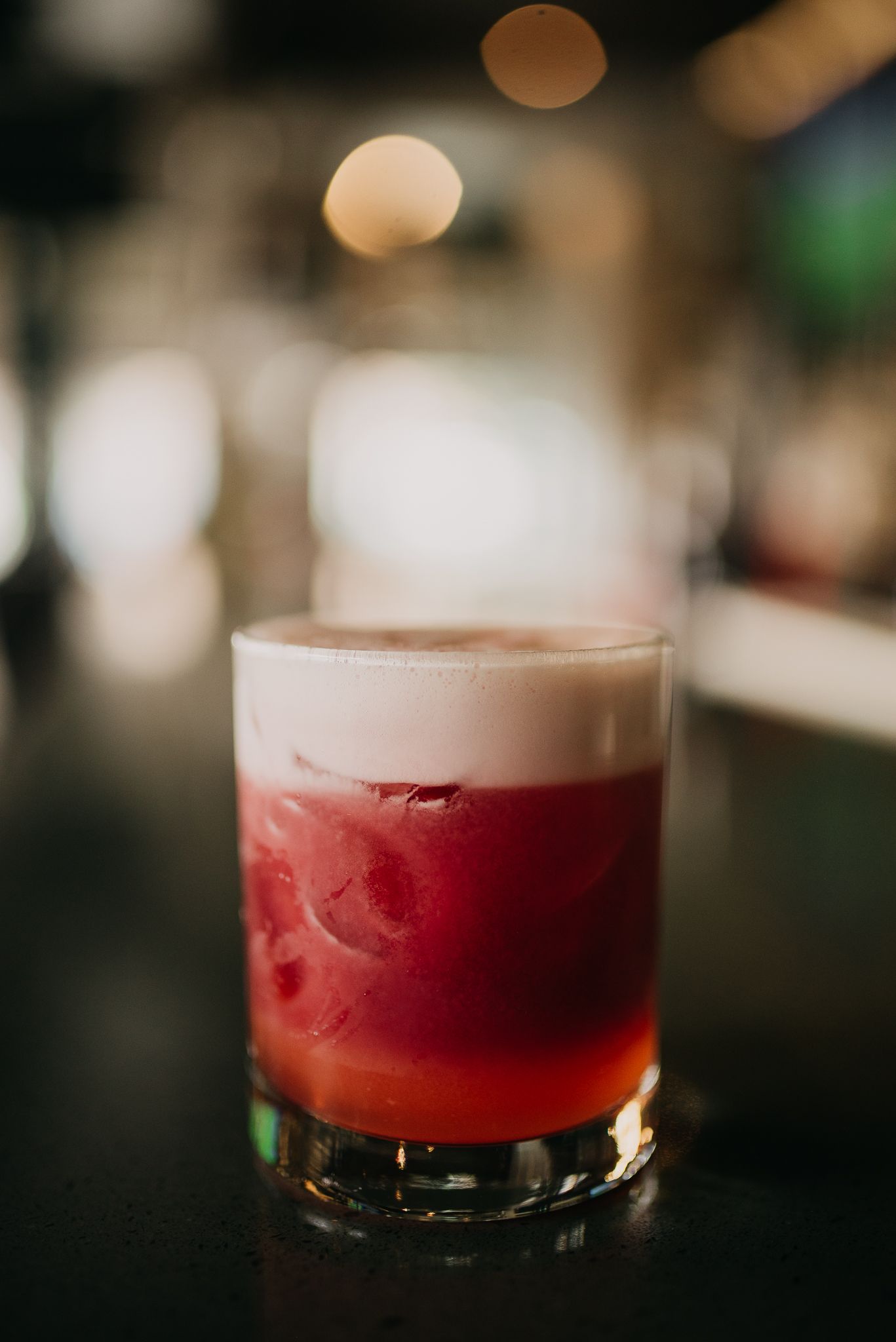 Red cocktail with white foam layer served in rocks glass on dark bar top.