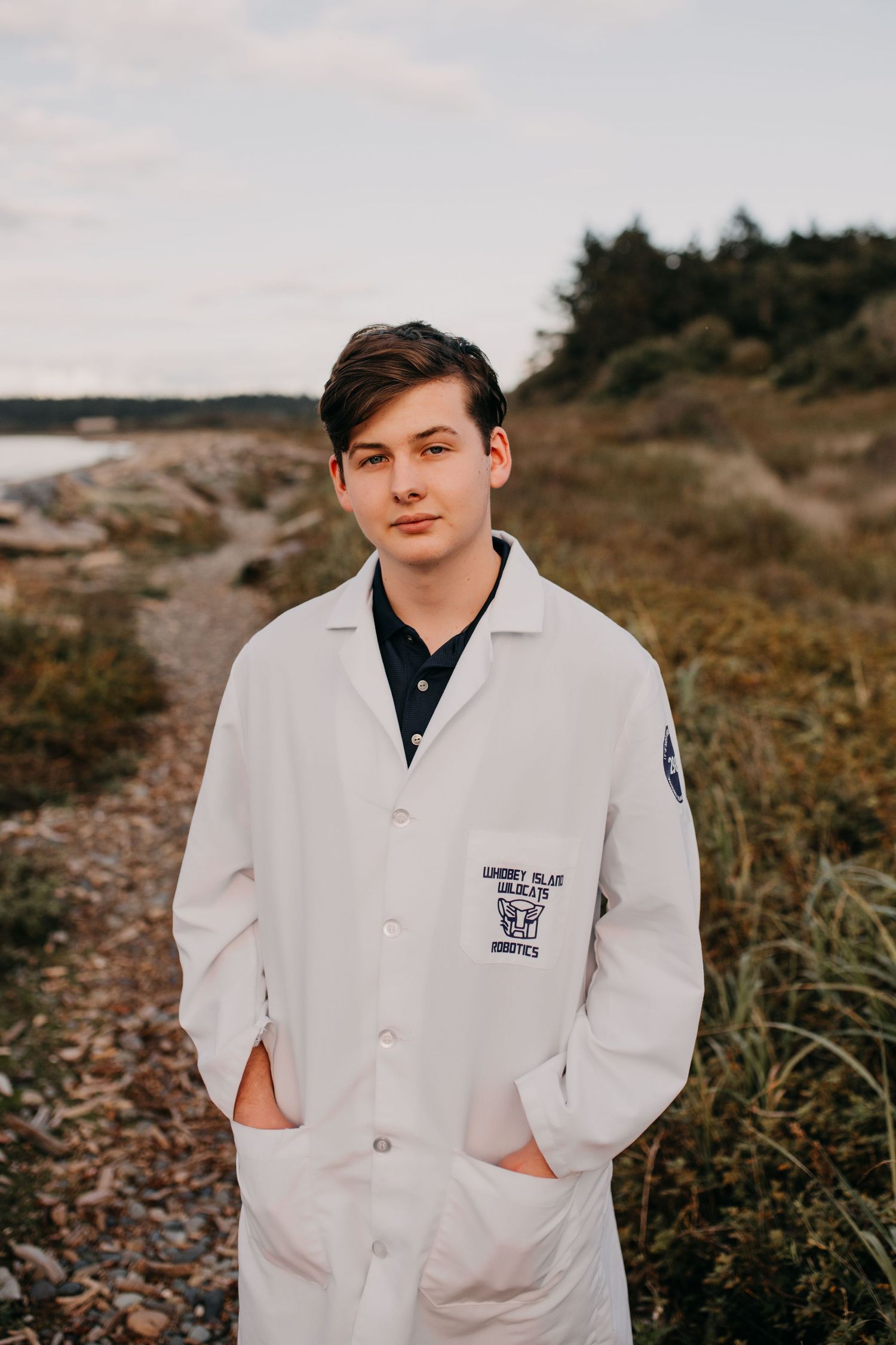 Young man in white lab coat stands on coastal trail with trees and water in background.