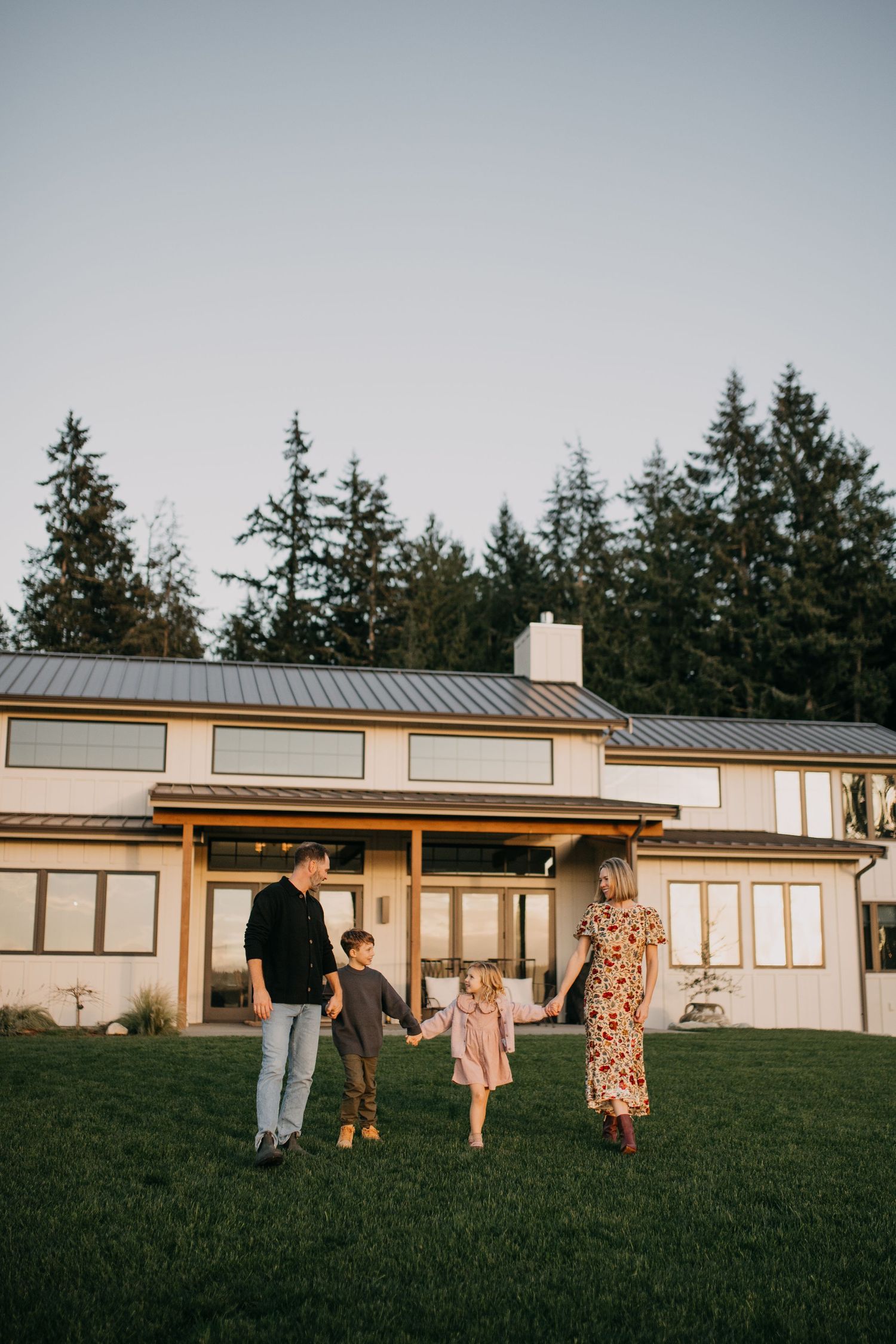 Family of four holding hands on green lawn in front of modern farmhouse surrounded by tall evergreen trees.