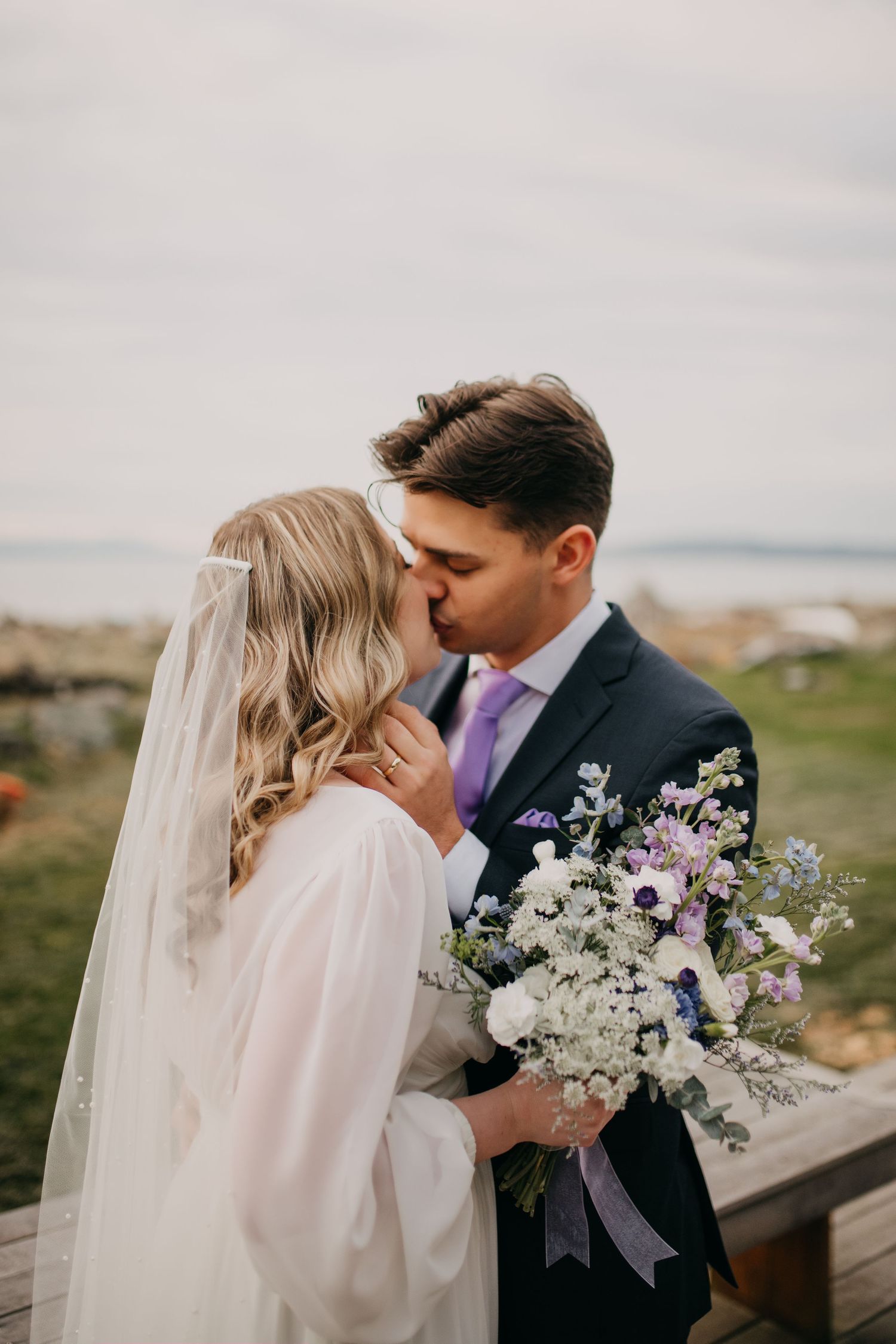 Bride and groom share a tender kiss while holding a wildflower bouquet at a coastal wedding.