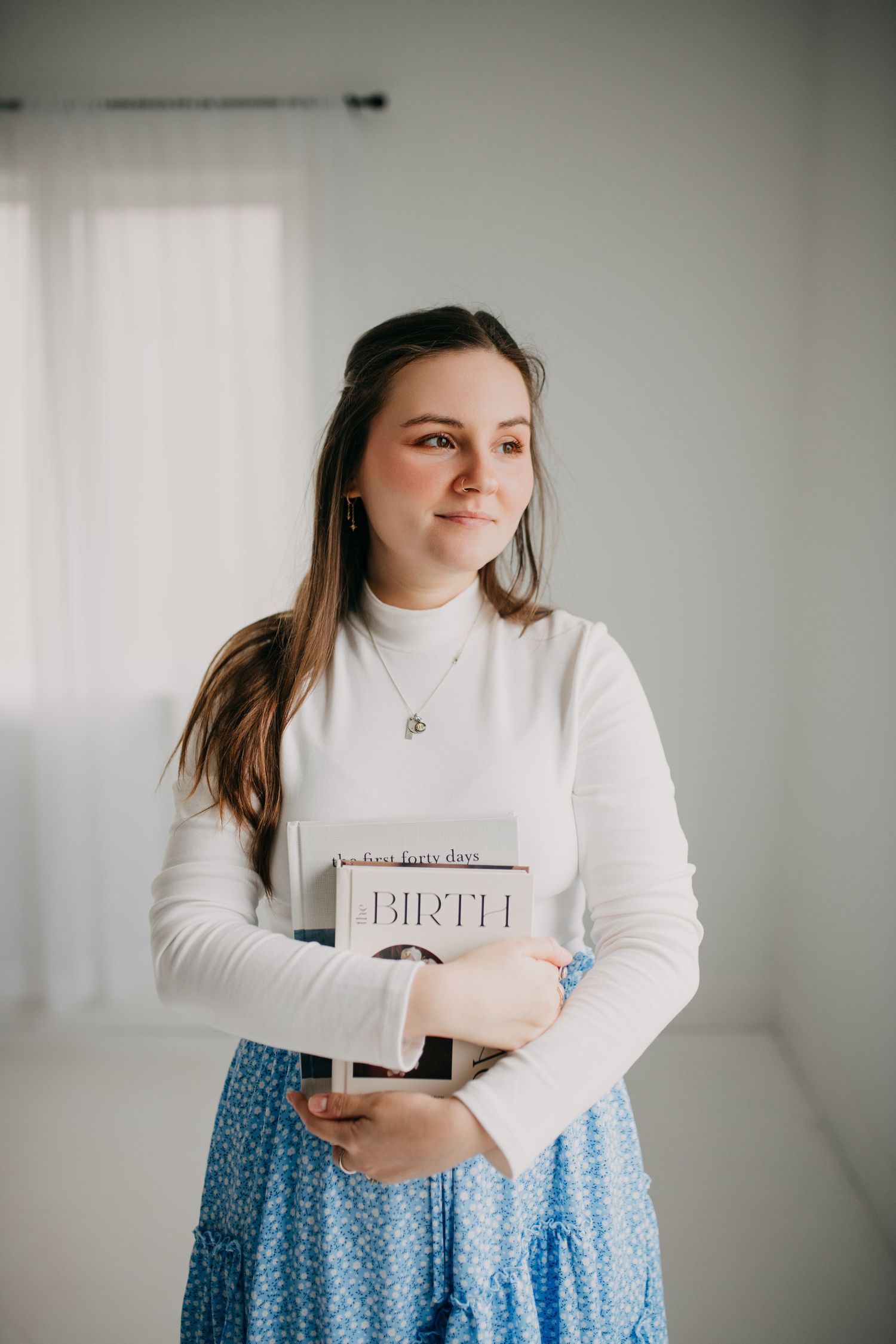 Young woman holding birth books smiling softly in a bright white room.