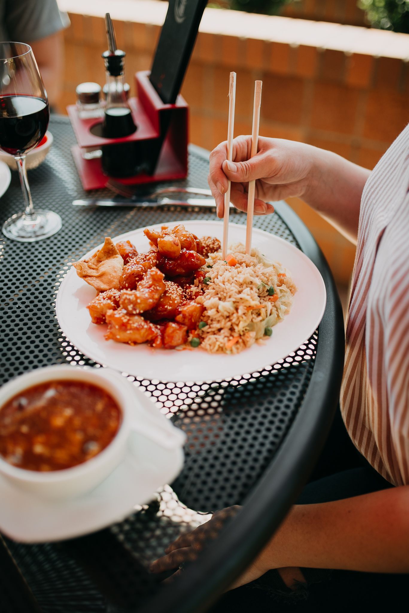 Person using chopsticks to eat glazed chicken with fried rice and dipping sauce outdoors.