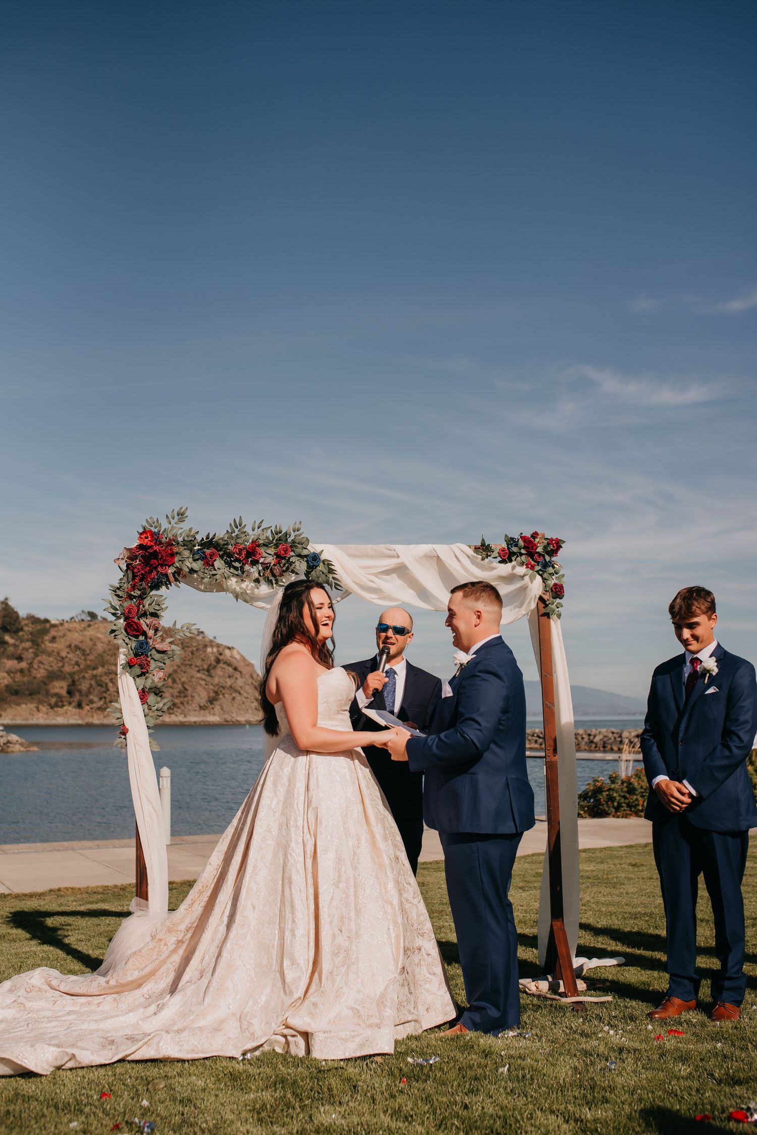 Bride and groom exchanging vows under floral arch with ocean and blue sky in background.