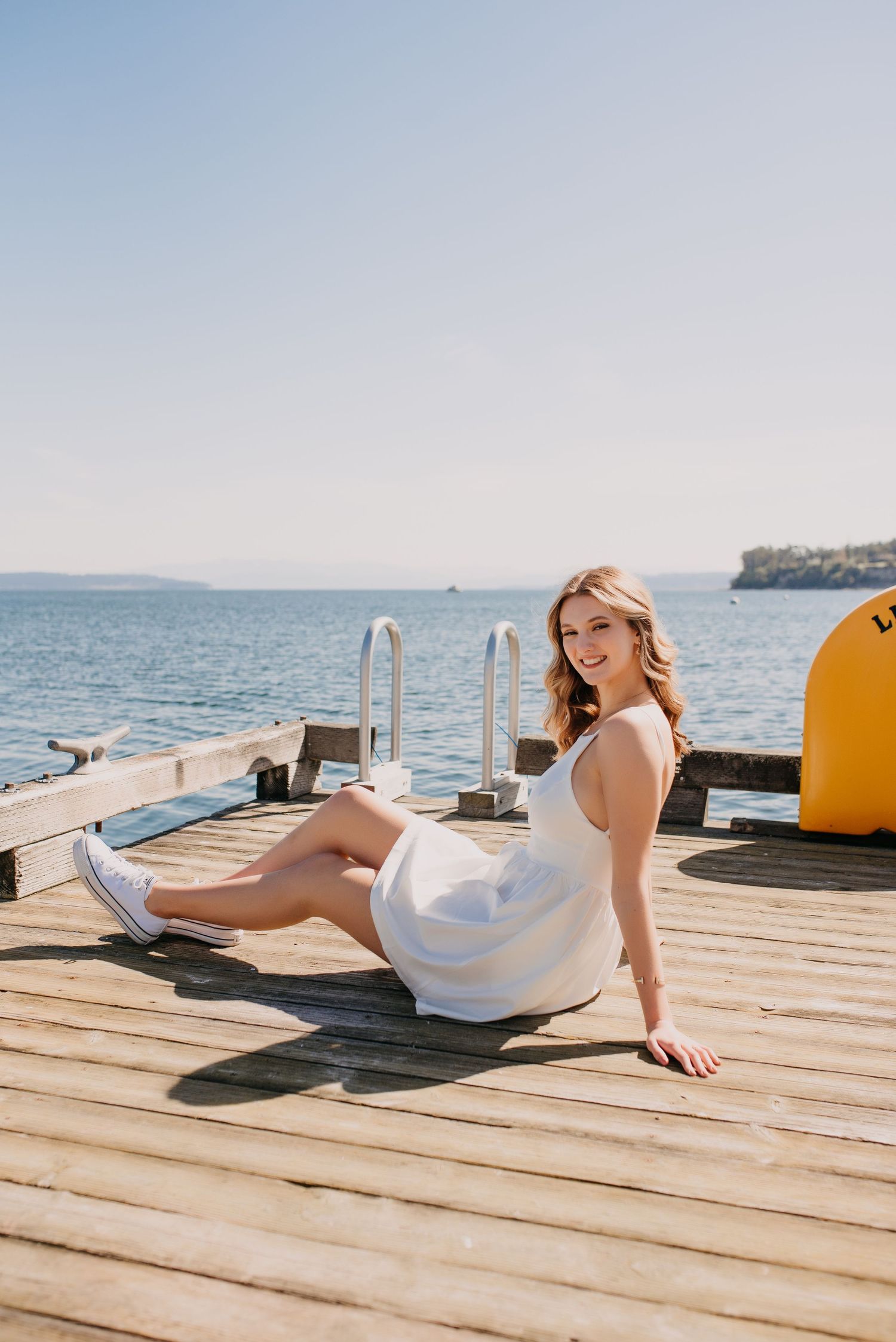 Smiling teen in white dress sits on wooden dock by blue ocean water on a clear day.