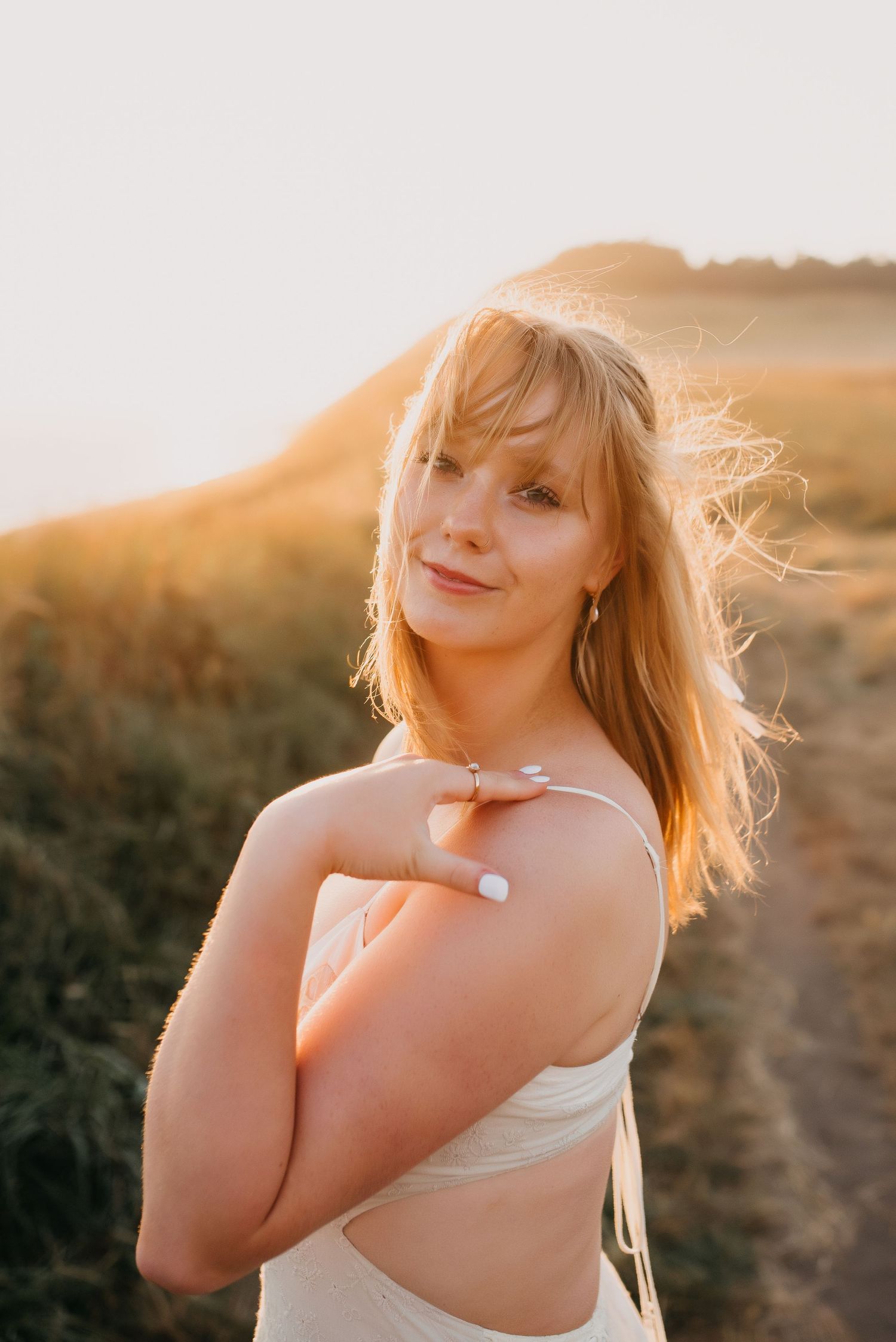Young blonde woman in white top posing outdoors during golden hour with wind in her hair.