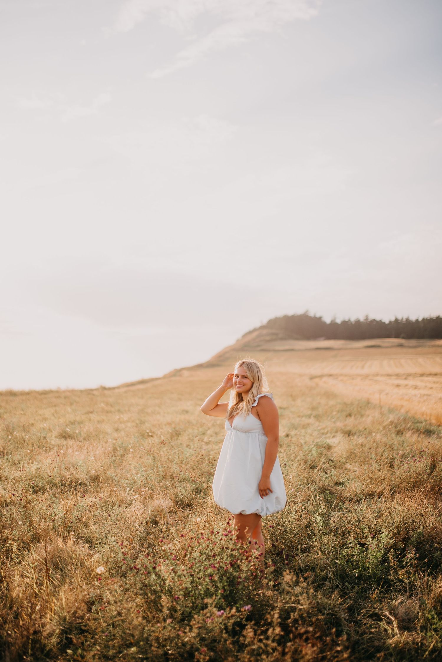 Blonde woman in white dress smiles while standing in a sunlit golden meadow.