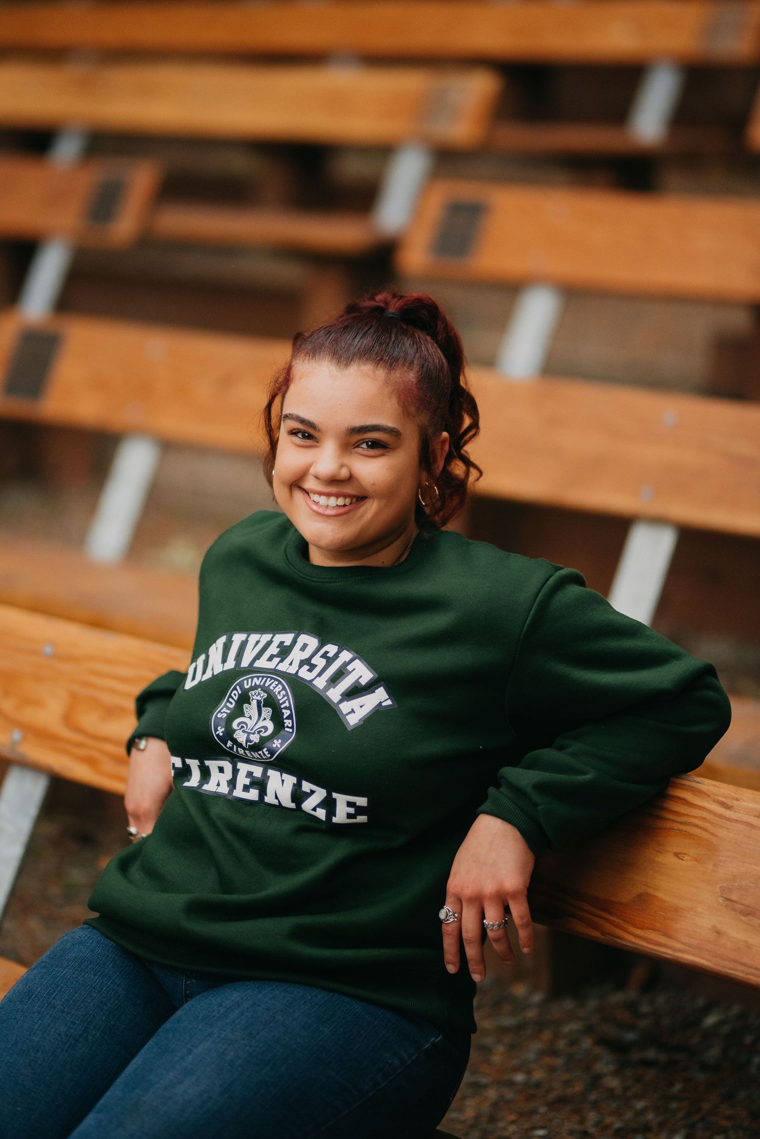 Smiling young woman in green Universita Firenze sweatshirt sits relaxed on wooden bleachers outdoors.