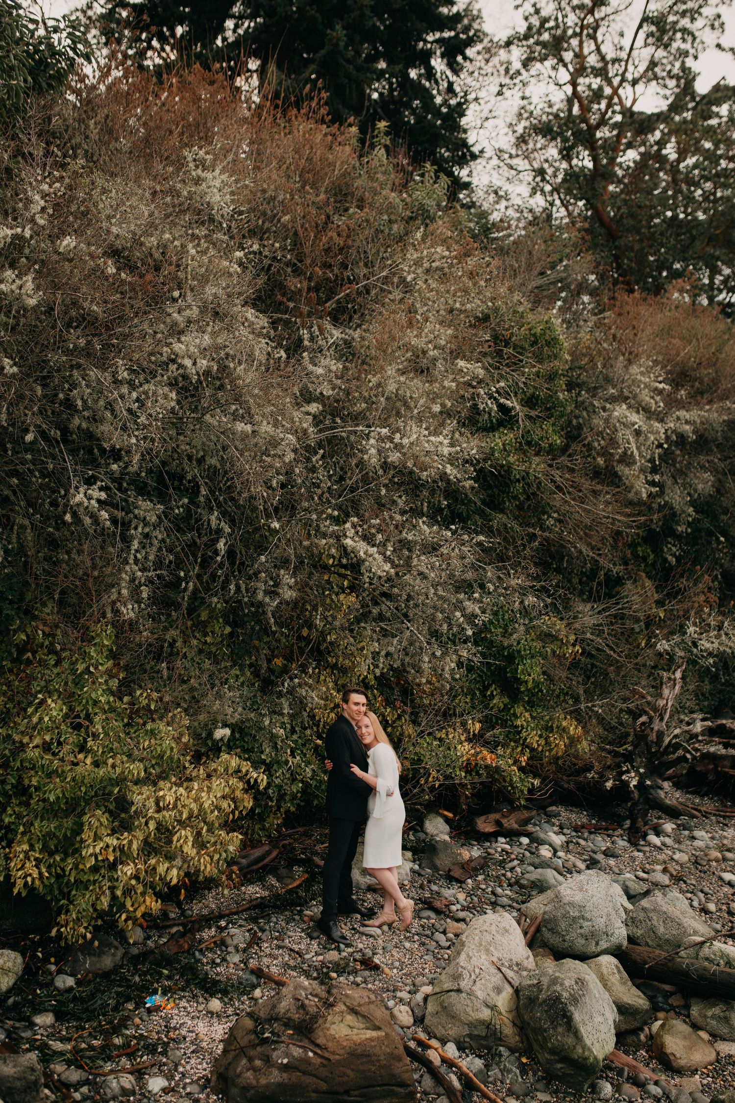 Couple embracing on rocky shoreline surrounded by lush autumn foliage and dense woodland trees.