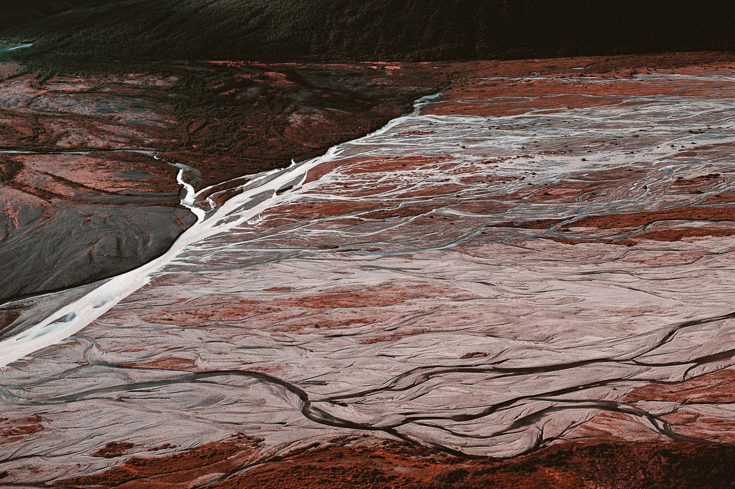 Aerial view of a glacier with red mineral deposits and braided meltwater streams below.