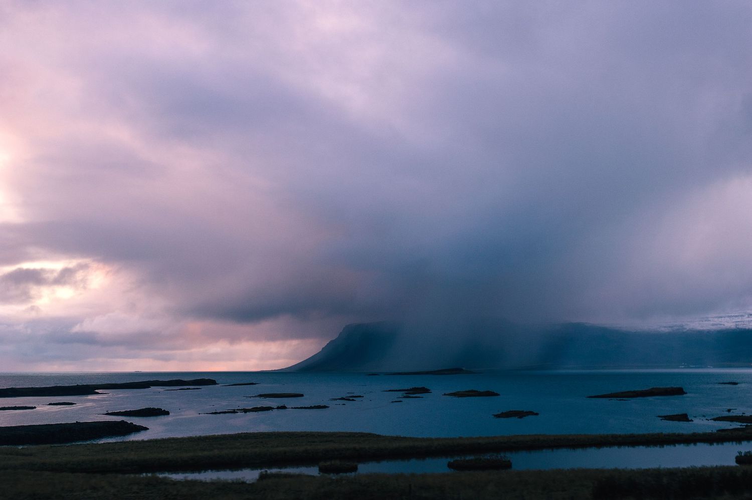 Stormy purple clouds descending over a volcanic island surrounded by shallow coastal waters in Iceland at twilight.