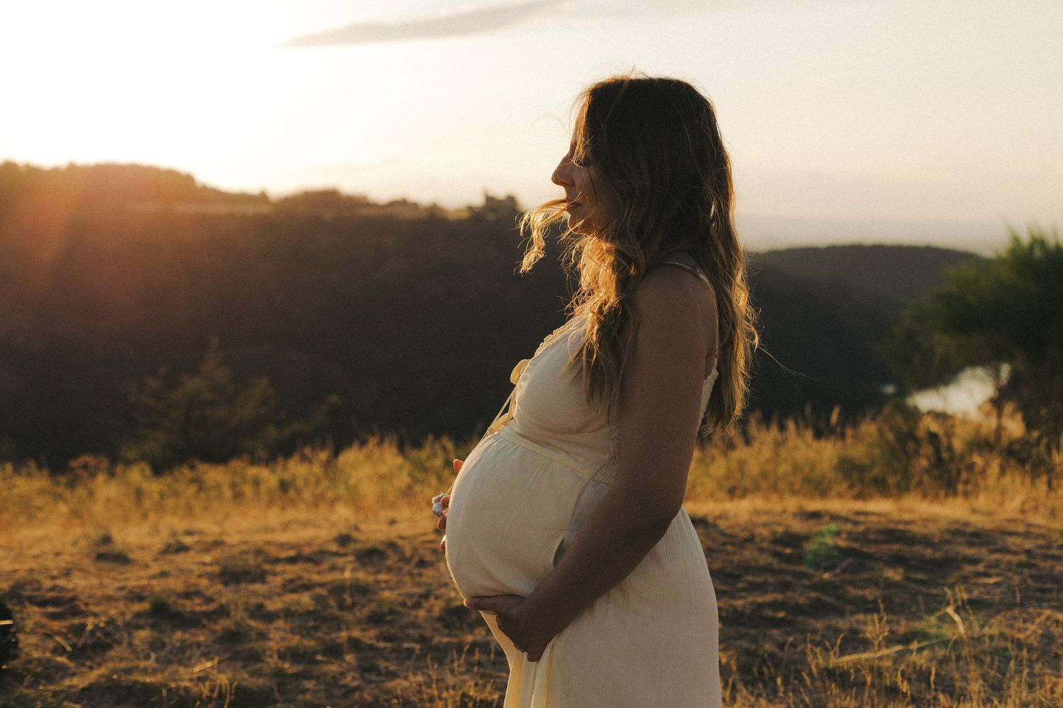 Pregnant woman in white dress holding her baby bump at golden hour sunset overlooking scenic hills.