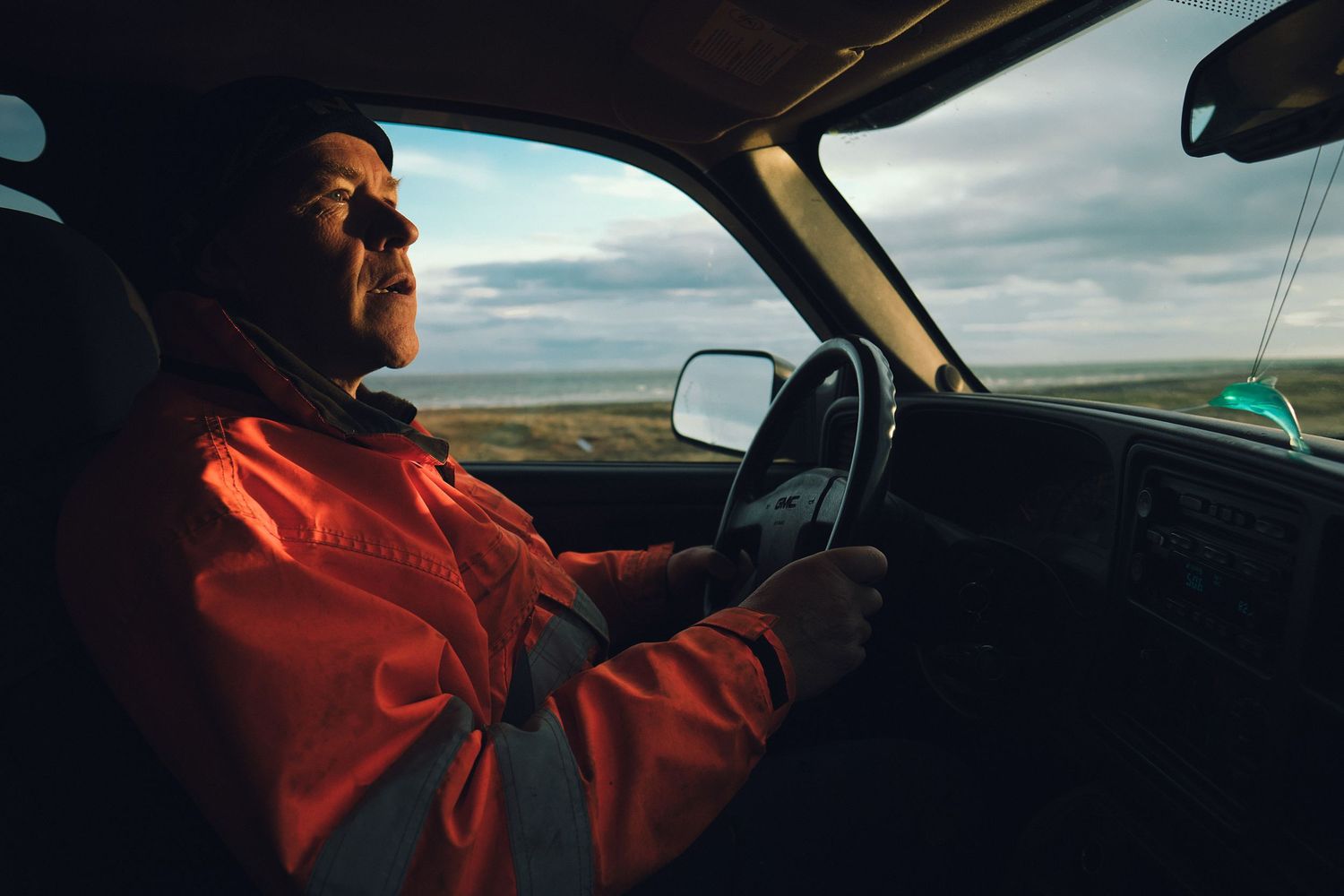 Man in a red jacket driving a vintage truck across the vast open Icelandic landscape under a cloudy sky.