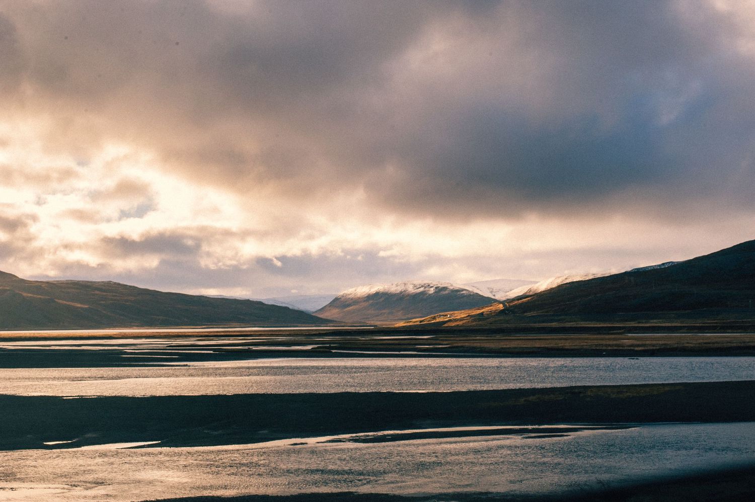 Dramatic sunset over a tranquil Icelandic fjord with snow-capped mountains and moody storm clouds reflected in calm waters.
