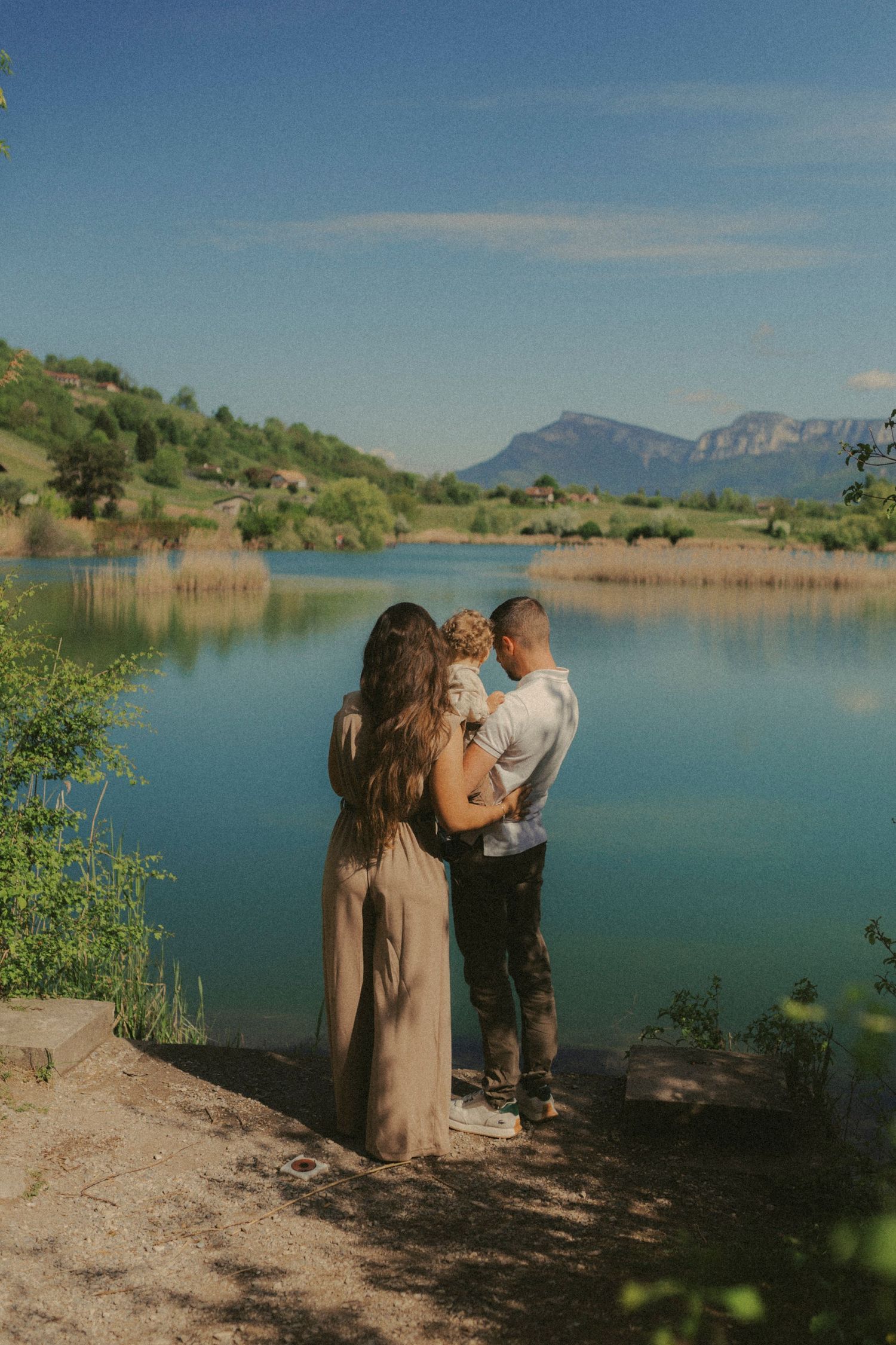 Family with a baby standing together by a serene mountain lake surrounded by lush green landscape.
