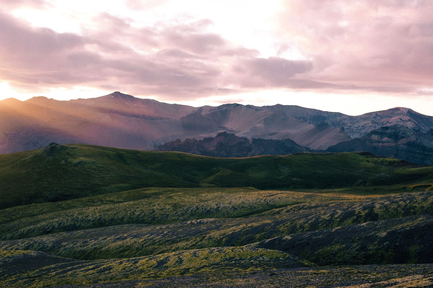 Golden sunset light breaks through clouds over rolling green volcanic highland hills.