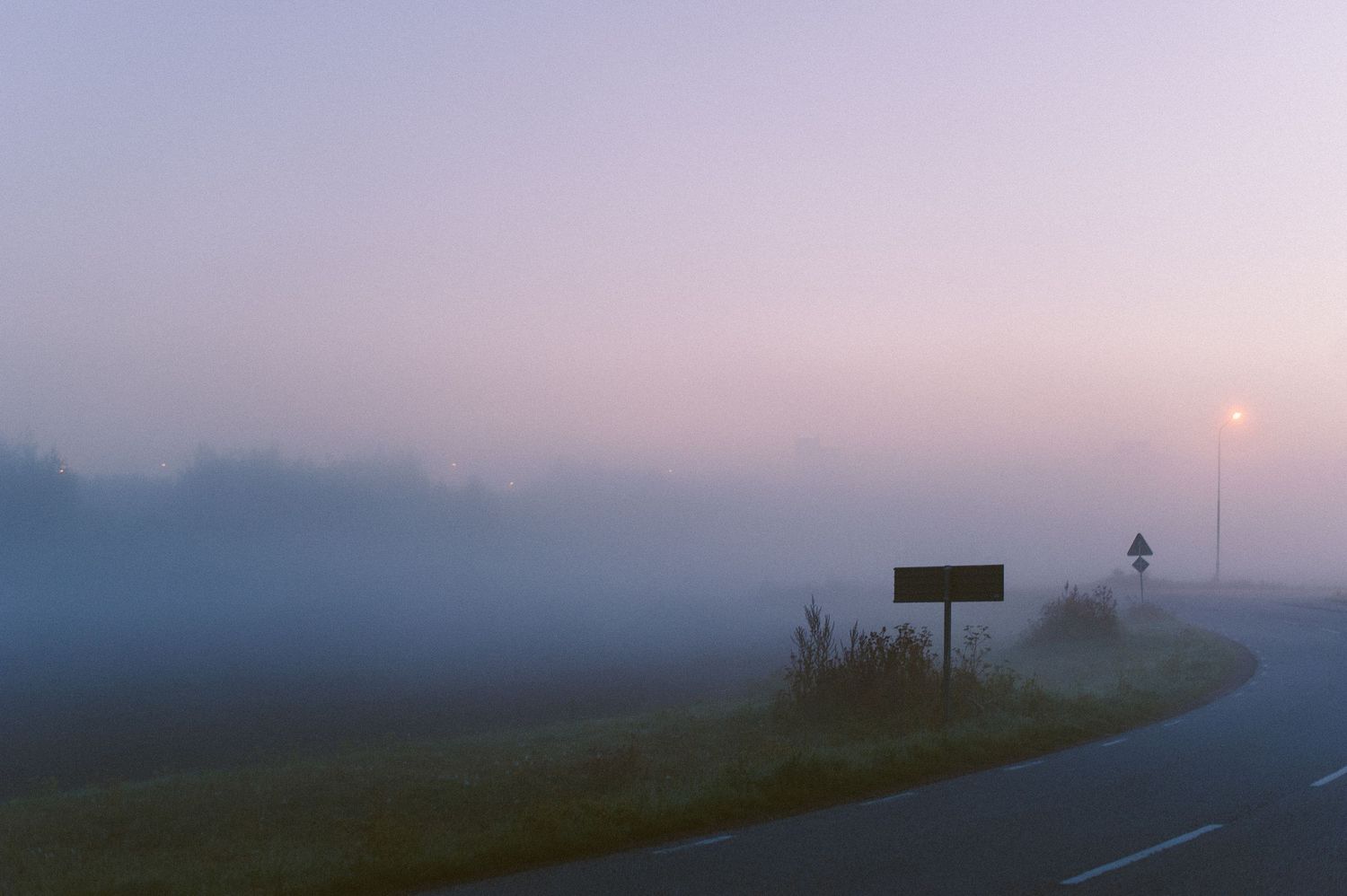 Misty foggy road at dawn with a roadside sign barely visible through the purple haze.