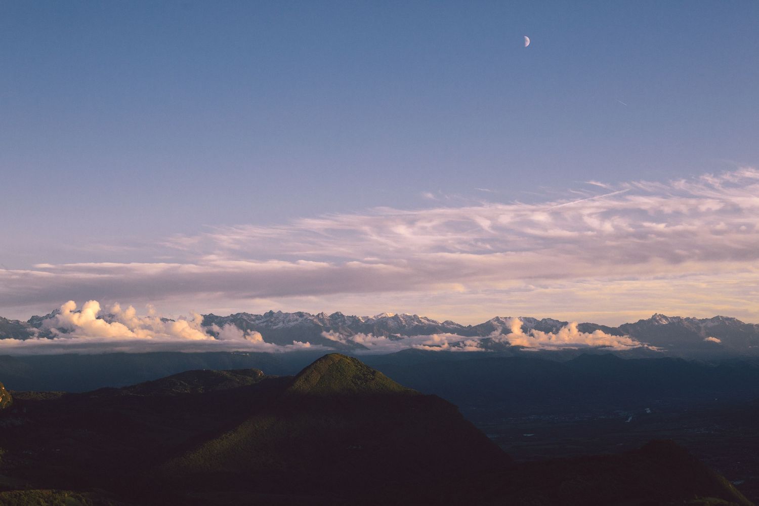 Panoramic mountain landscape at dusk with layered clouds and a crescent moon in a soft purple sky.