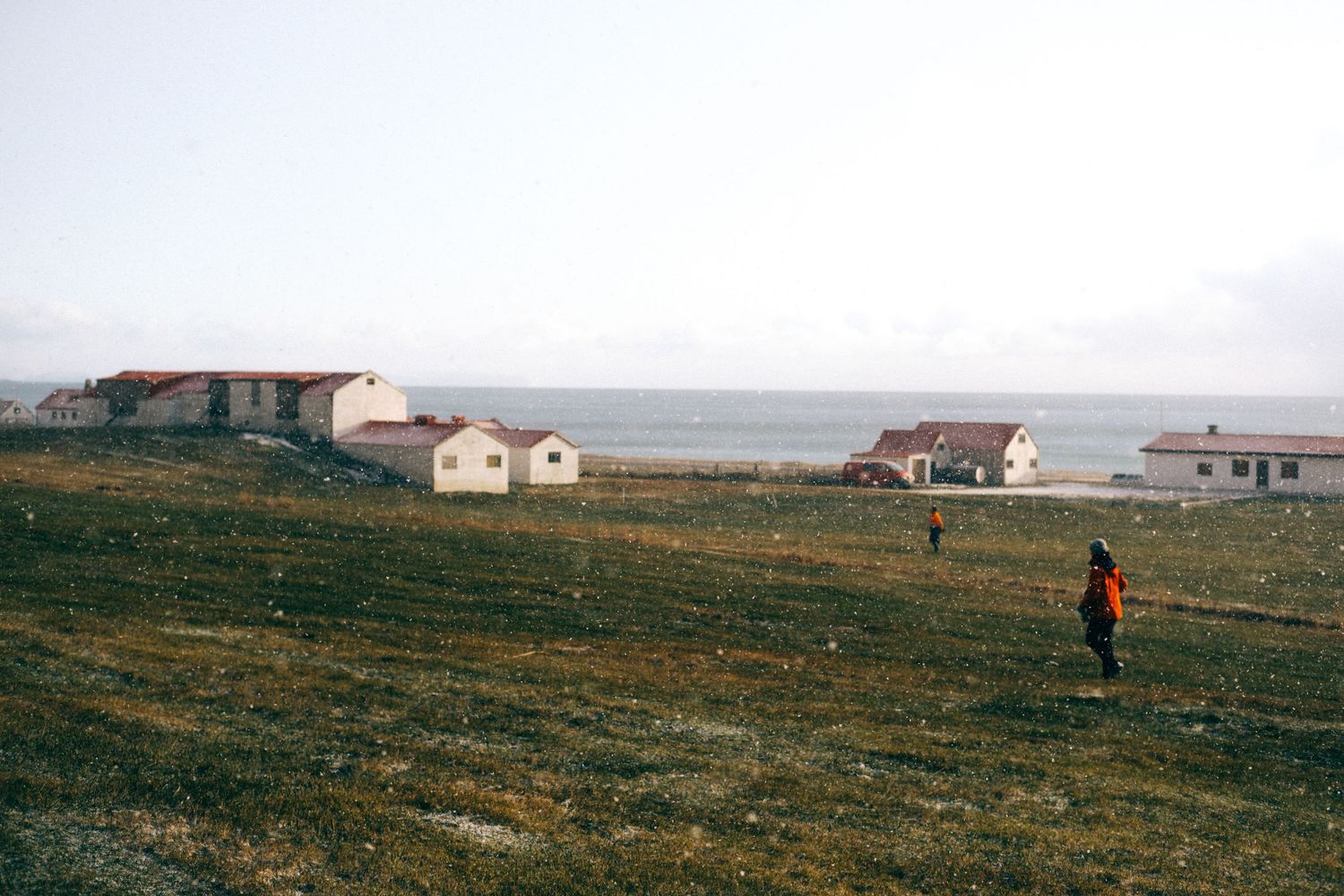 Person walking across a vast green coastal landscape with small houses and ocean views in the background.