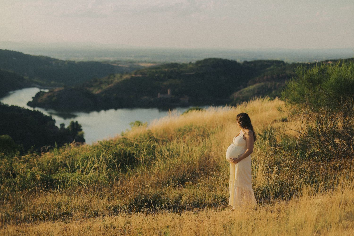 Pregnant woman in a white dress standing on a hillside overlooking a scenic river valley at sunset.