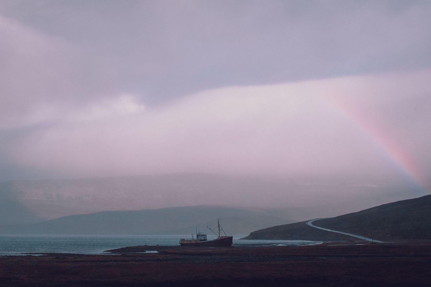 A rainbow arcs over a misty fjord with an abandoned shipwreck on the rocky shoreline below.