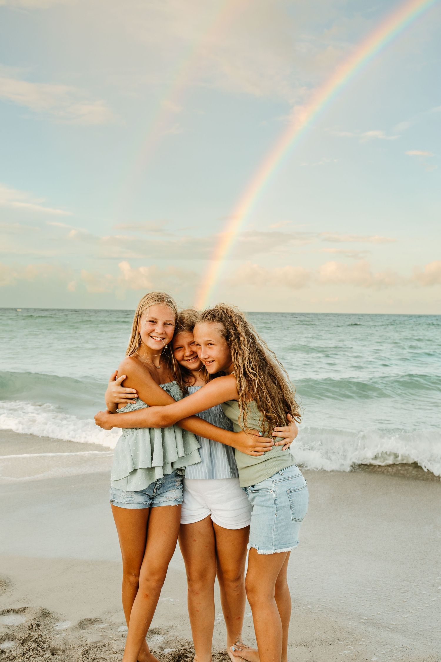 Three friends embrace while standing on a beach at sunset with a rainbow visible in the sky.