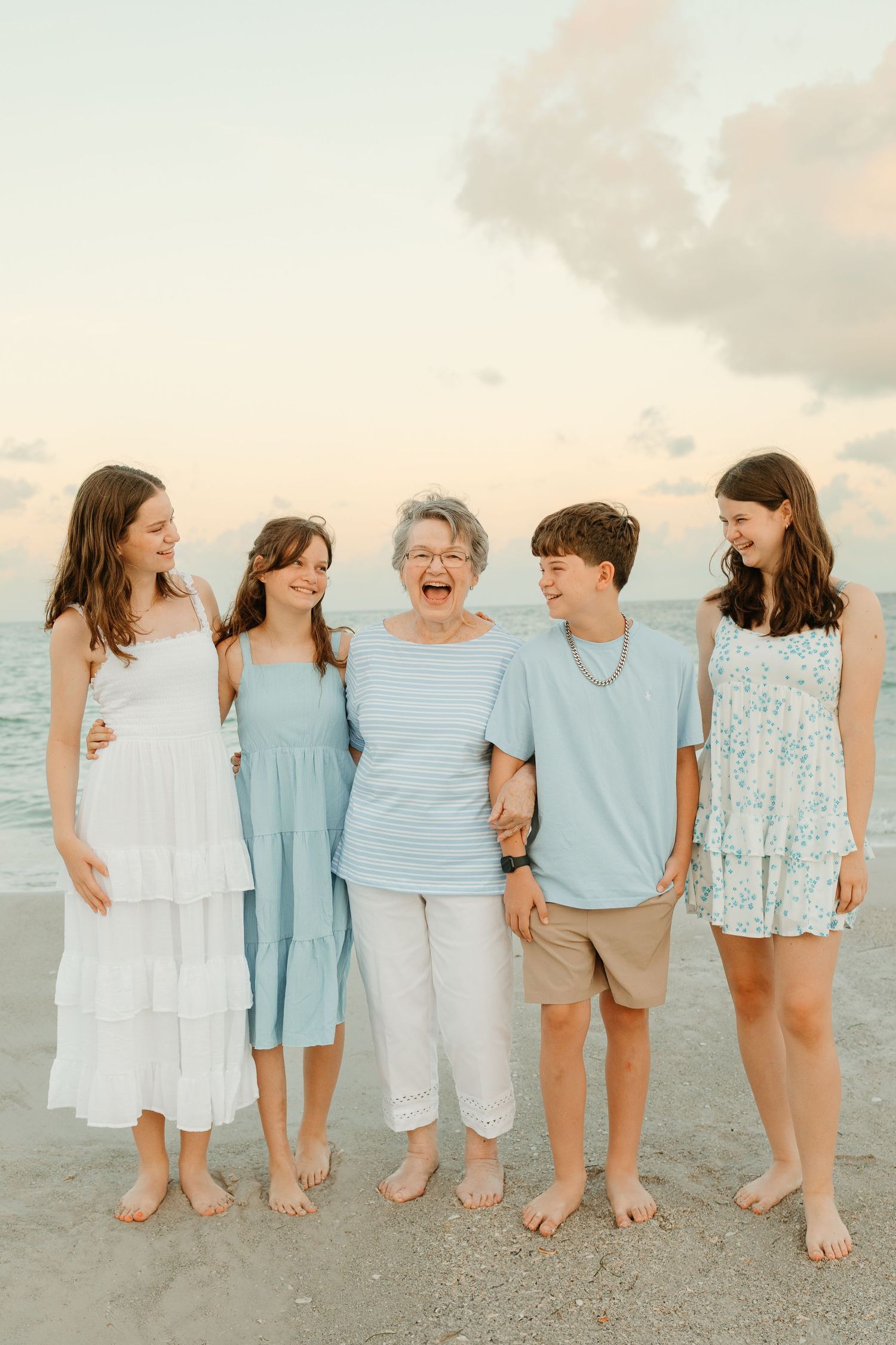 A group of people in light blue and white outfits stand together on a beach at sunset for a family photo.