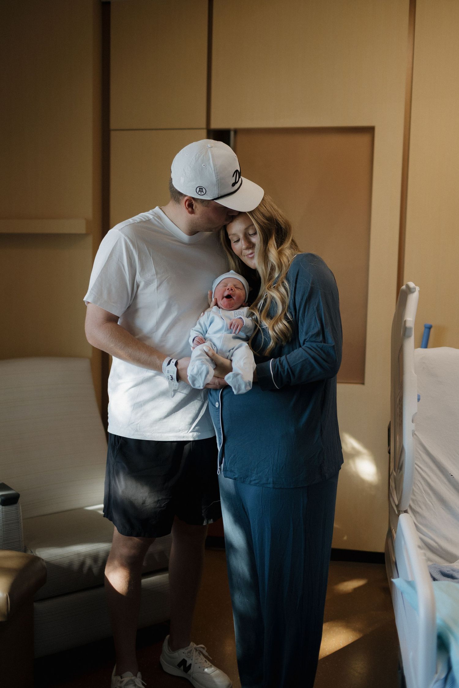 New parents hold their newborn baby together in a hospital room with warm lighting and medical equipment visible.