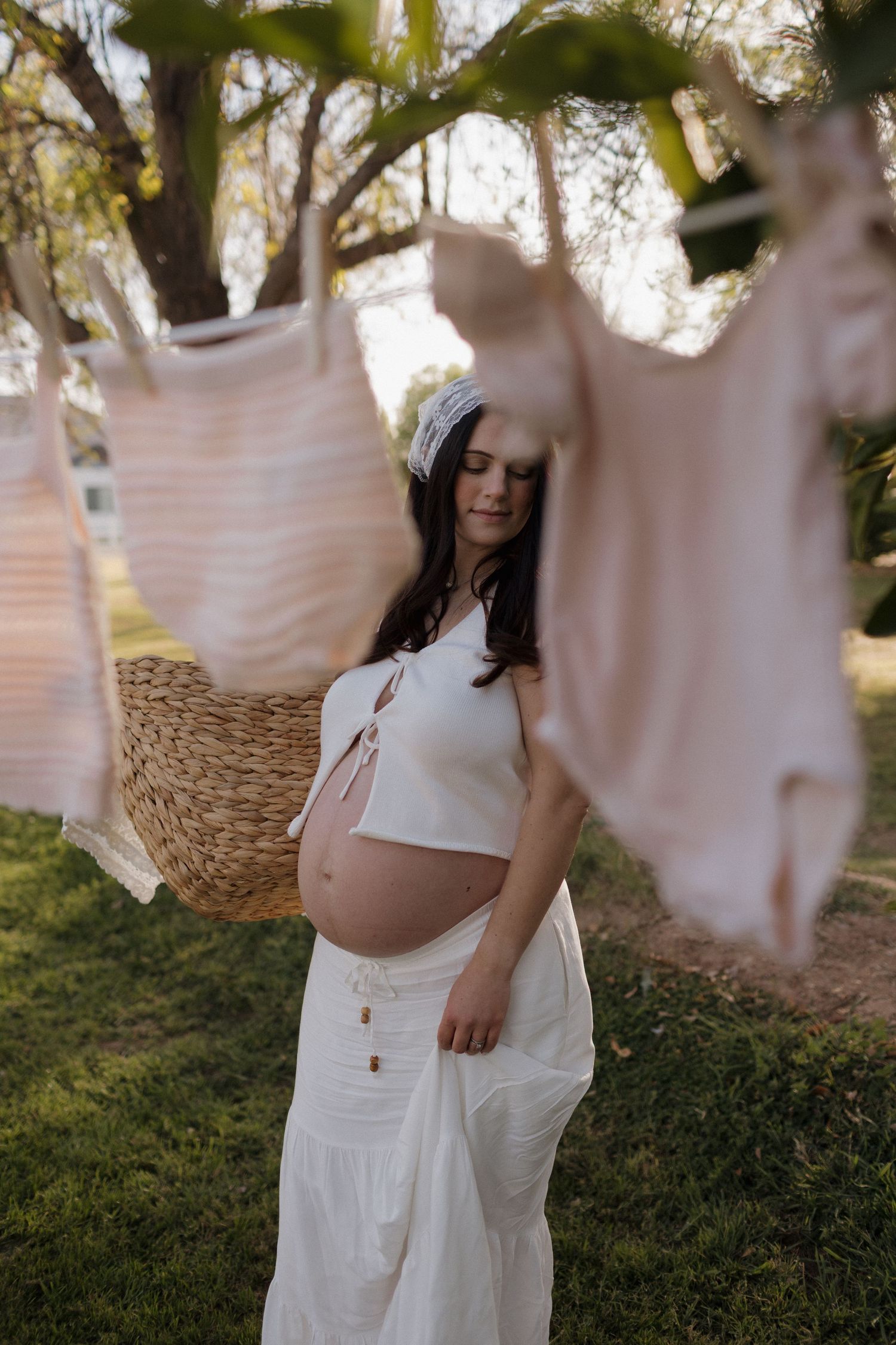 Pregnant woman in white crop top and pants stands outdoors near clothesline with pink baby clothes hanging in soft light.