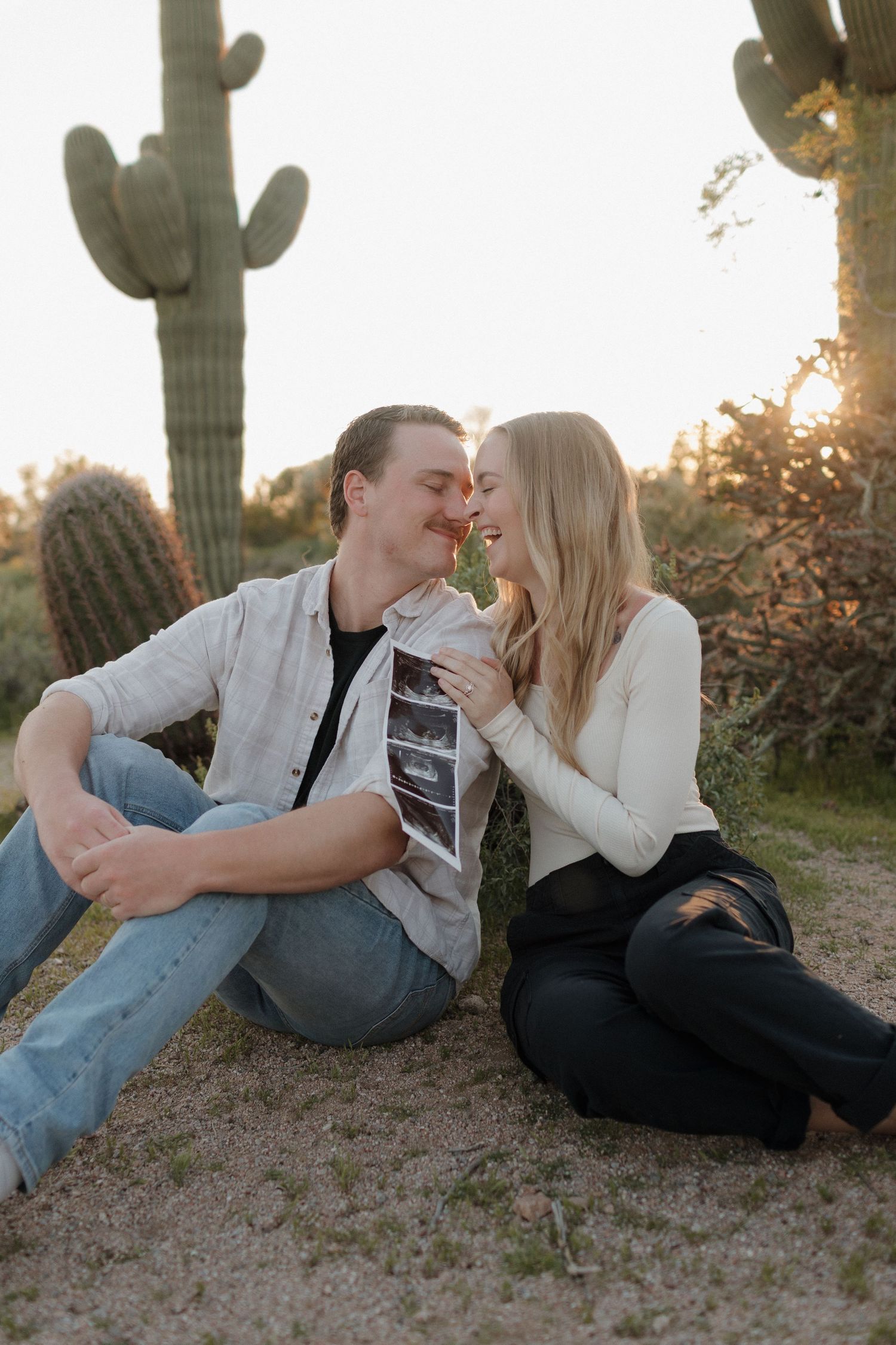 Desert maternity announcement photo with couple holding sonogram against cactus backdrop.