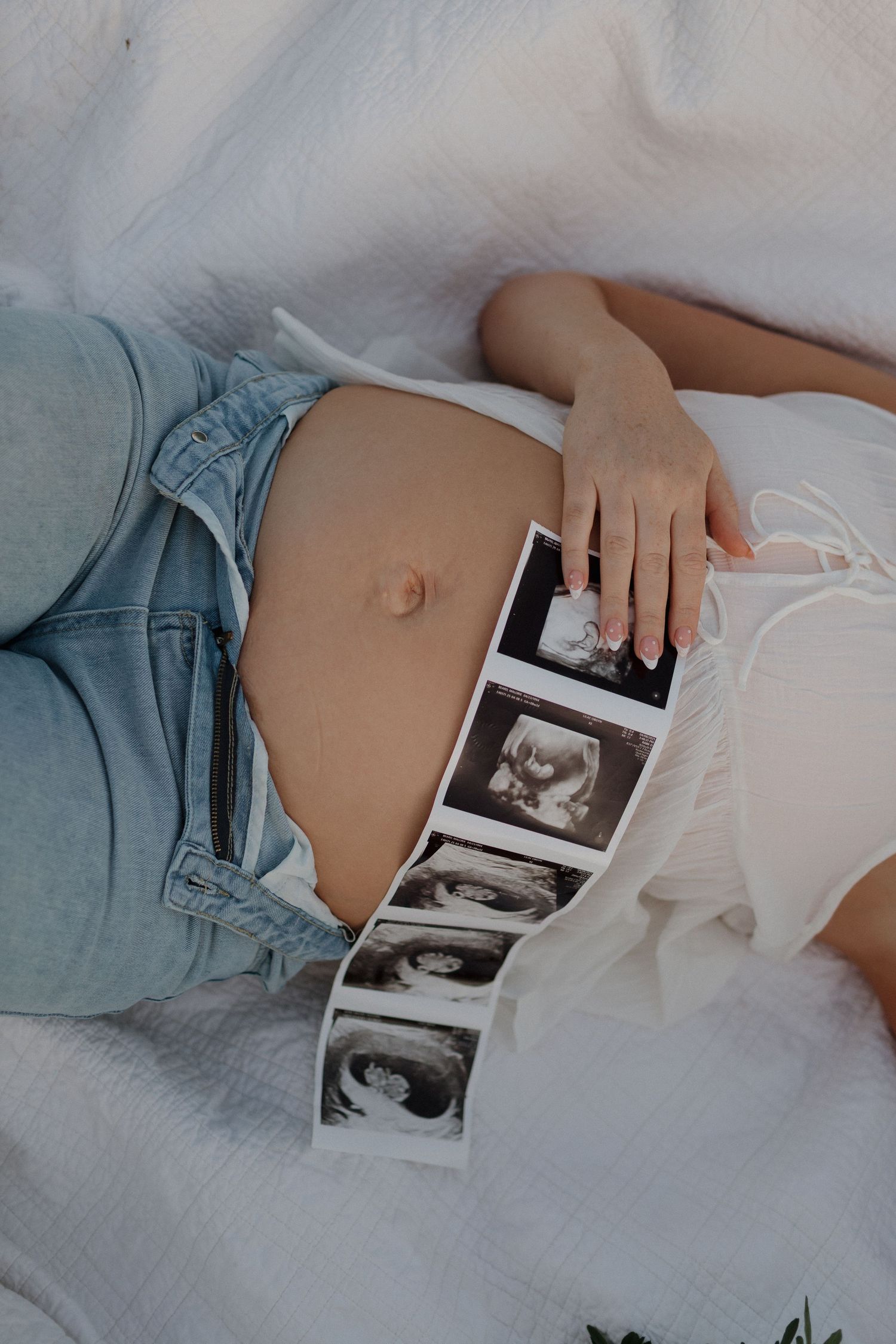 Pregnant woman in denim shorts lying down holding ultrasound photos on white blanket