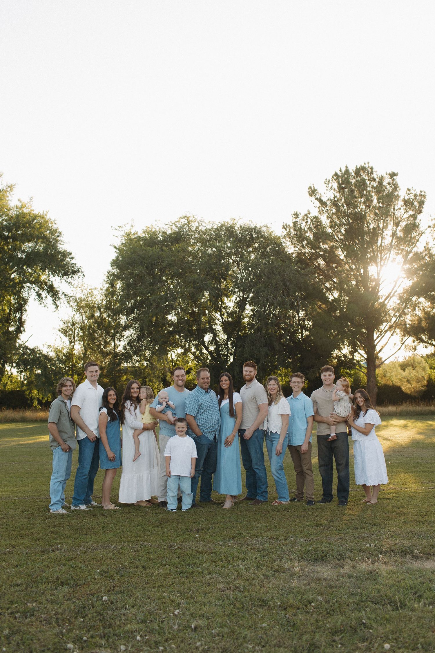 Large family group portrait outdoors at golden hour, wearing coordinated blue and white outfits, posing on green grass with trees behind.