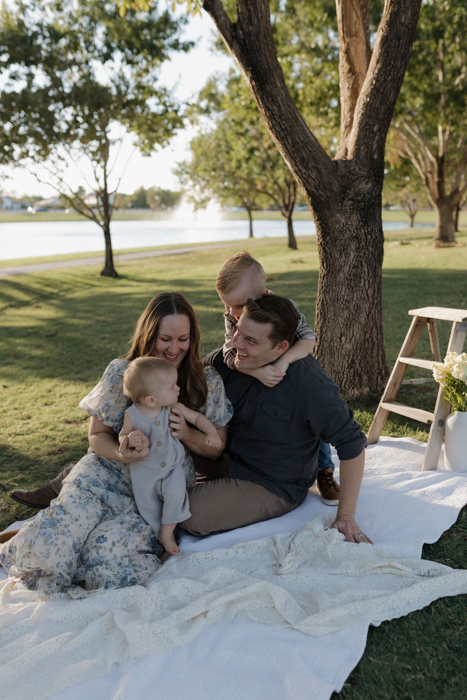 A happy family of four enjoys a sunny outdoor picnic on a white blanket near a tree by a lake.