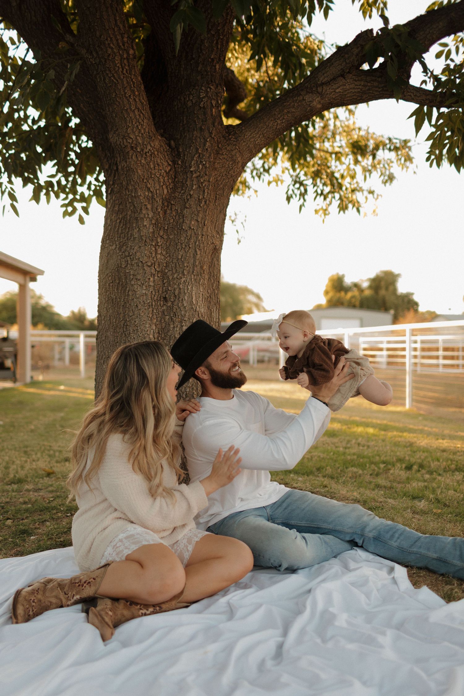 Couple having a relaxing picnic under large tree on white blanket at sunset near farm with wooden fences.