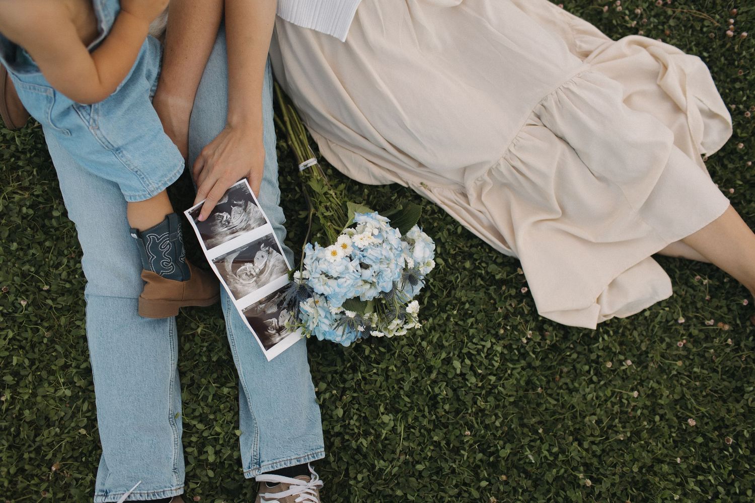 Couple sitting on grass holding ultrasound photos with blue hydrangea bouquet, pregnancy announcement outdoor photo.