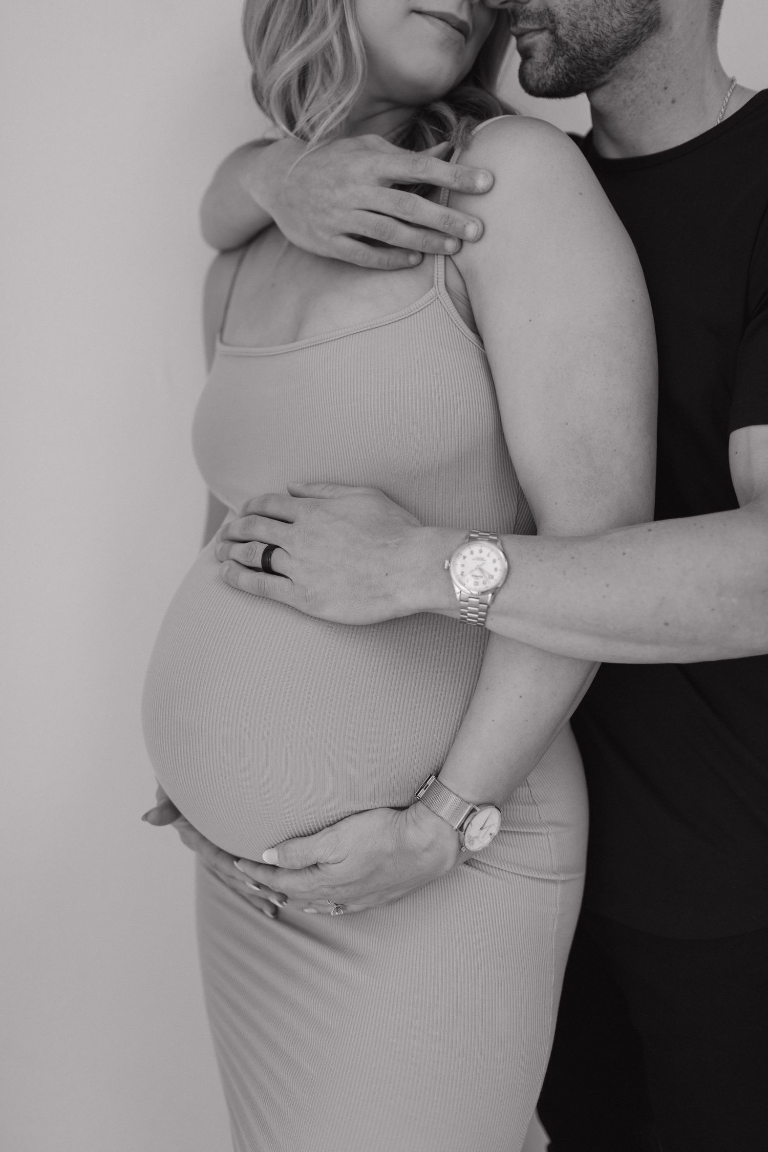 Black and white maternity photo showing a couple embracing with hands tenderly placed on pregnant belly, wearing casual attire.