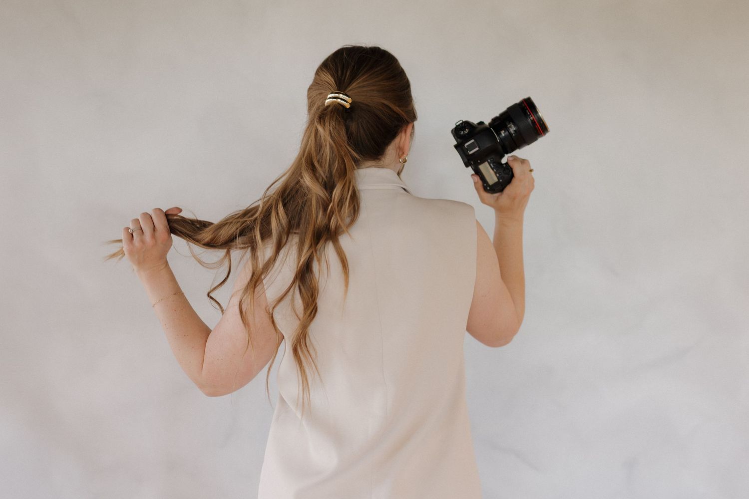 Photographer with long wavy brown hair holds a camera while standing against a light gray backdrop, viewed from behind.